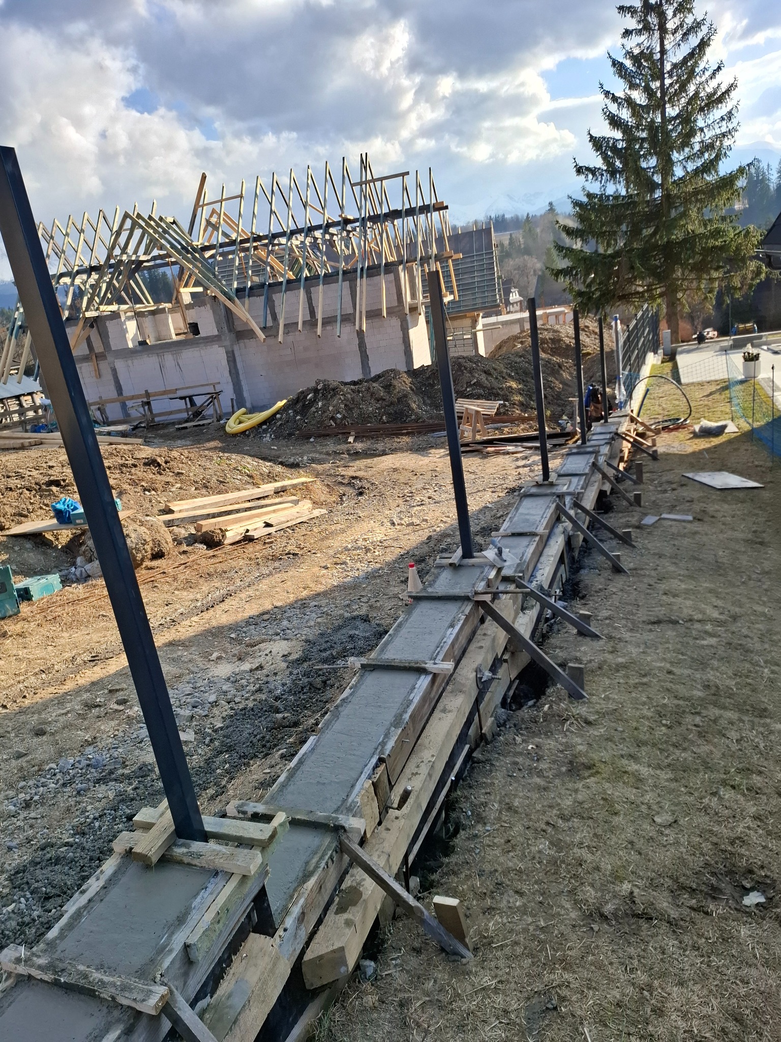 Fundament w szalunku, zalany betonem, z wystającymi stalowymi słupami. W tle budowa domu w stanie surowym otwartym i drzewo iglaste.