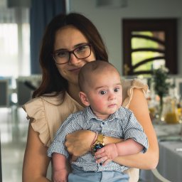 JMWieczorek - Kobieta w okularach trzyma na rękach niemowlę ubrane w koszulę w kratę i niebieskie spodnie. W tle widoczny stół z dekoracjami.