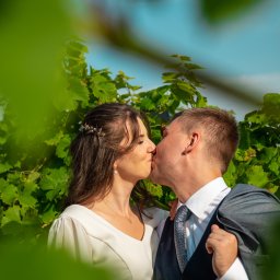 JMWieczorek - Para młoda w plenerze, pocałunek na tle zielonych liści. Panna młoda w białej sukni, pan młody w garniturze z marynarką na ramieniu. Romantyczna sesja.