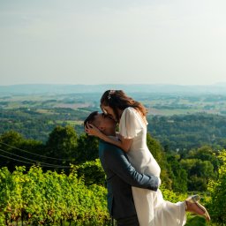 JMWieczorek - Para młoda w objęciach na tle winnicy i rozległego krajobrazu. Panna młoda w białej sukni, pan młody w szarym garniturze. Ujęcie z dołu, podkreślające radość chwili.