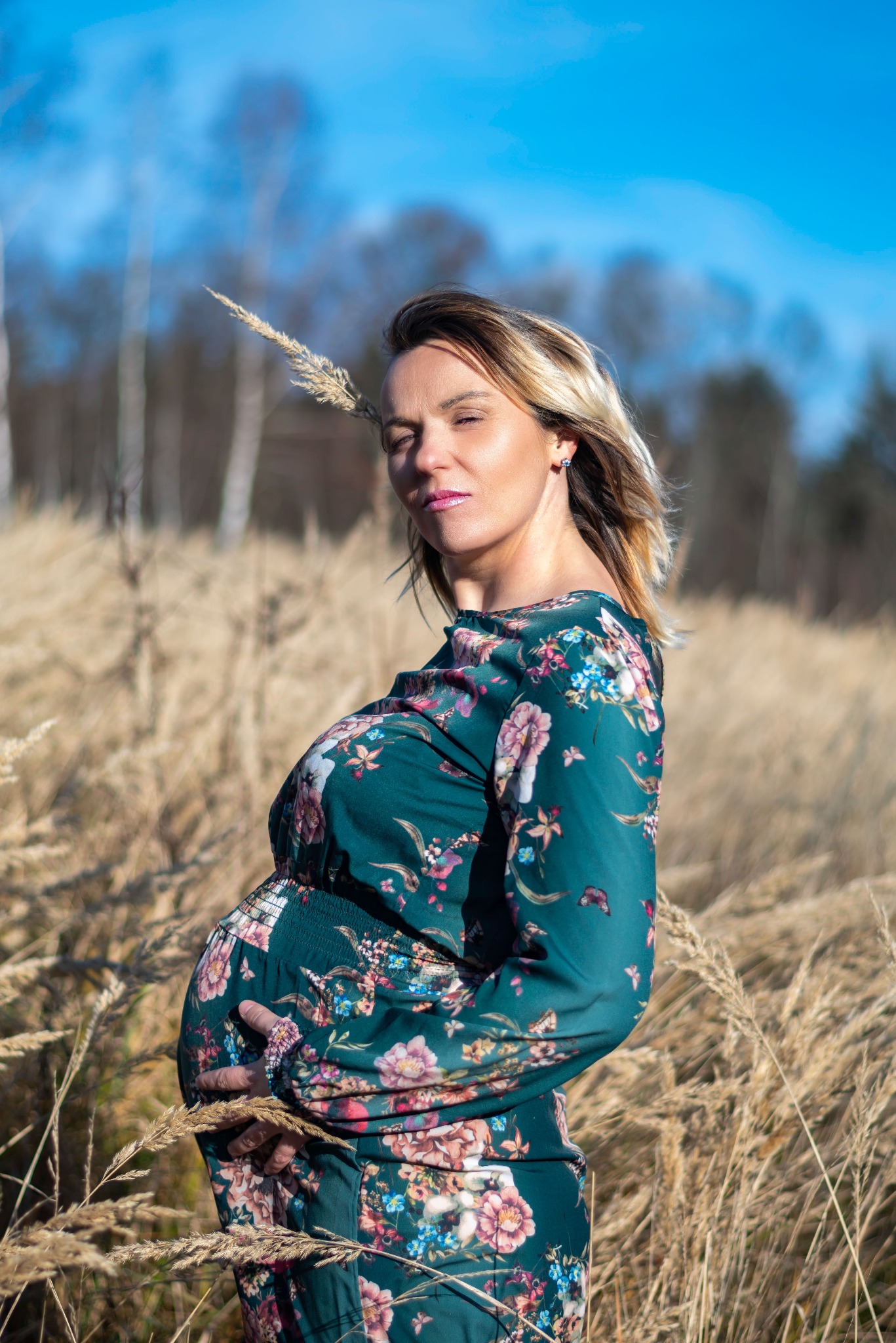 Kobieta w ciąży w kwiecistej sukience, pozująca na tle łąki z wysoką trawą i błękitnego nieba. Sesja ciążowa w naturalnym otoczeniu, delikatne oświetlenie.