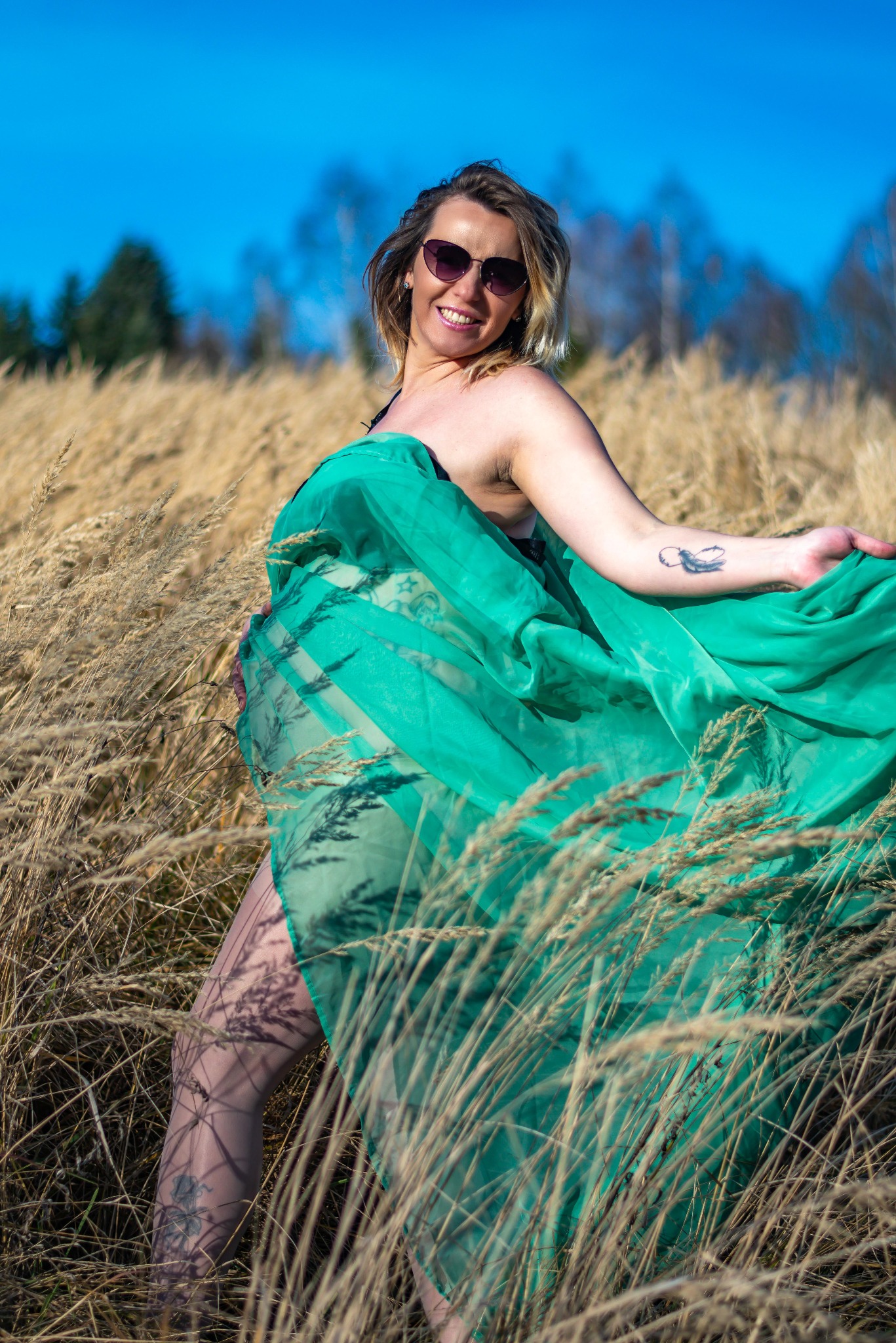 Kobieta w okularach i zwiewnej, zielonej tkaninie pozuje w polu trzcin, z tatuażem na ramieniu, w słoneczny dzień. Uśmiech, naturalne światło, niebieskie niebo w tle.