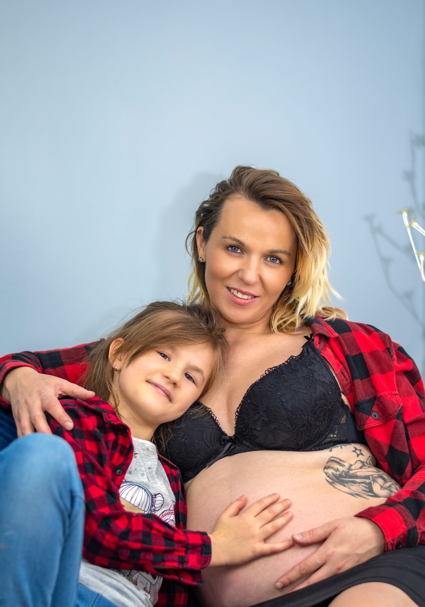 Portret uśmiechniętej, ciężarnej kobiety w czarnym biustonoszu i czerwonej koszuli w kratę, obejmującej córkę. Dziecko trzyma rękę na brzuchu matki.