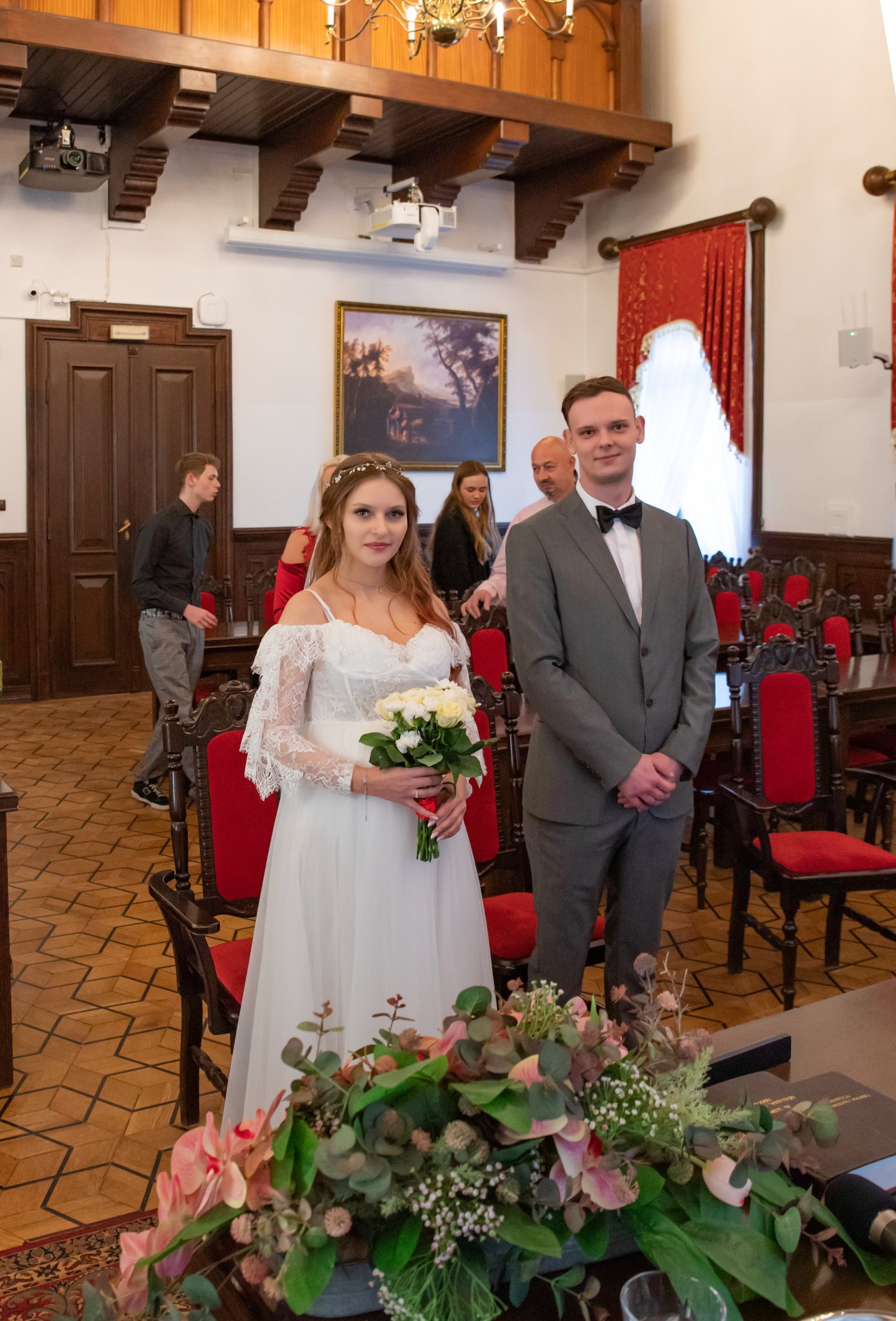 Panna młoda w koronkowej sukni trzyma bukiet, obok pan młody w szarym garniturze z muchą, w tle goście i dekoracje sali ślubów z drewnianym stropem.