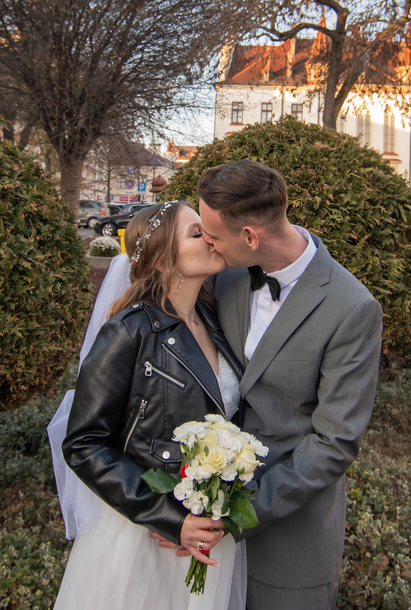 Pocałunek pary młodej w plenerze: panna młoda w sukni i ramonesce, pan młody w szarym garniturze. W tle budynek z czerwonym dachem i zielone krzewy.