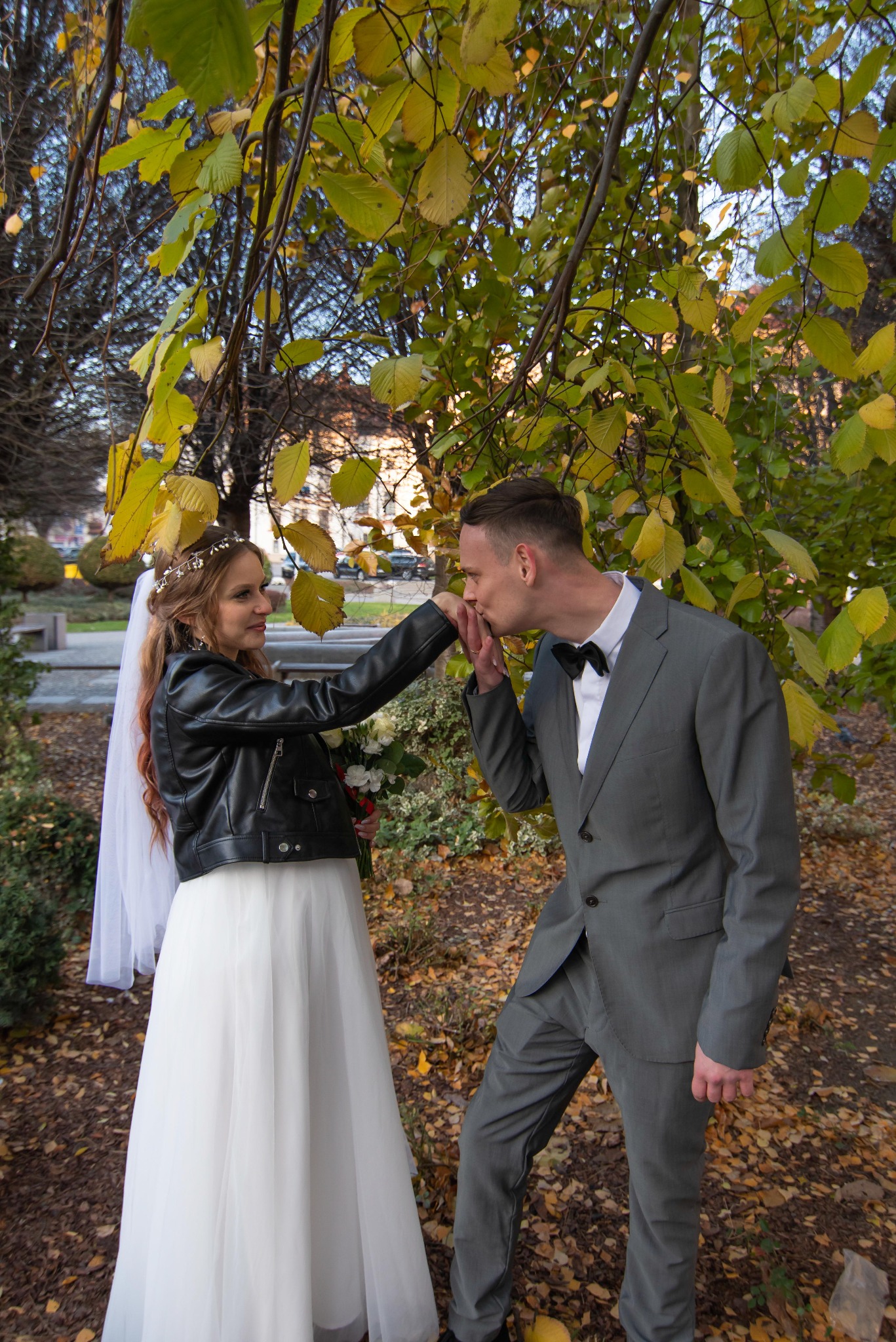 Jesienna sesja ślubna w parku: Panna młoda w skórzanej kurtce i welonie, pan młody w szarym garniturze całuje jej dłoń pod drzewem z żółtymi liśćmi.