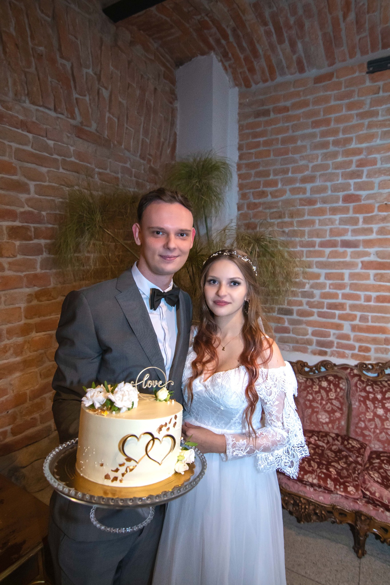 Para młoda z tortem weselnym na tle ceglanej ściany i dekoracyjnej sofy. Panna młoda w koronkowej sukni, pan młody w szarym garniturze. Tort zdobiony sercami i napisem 'love'.