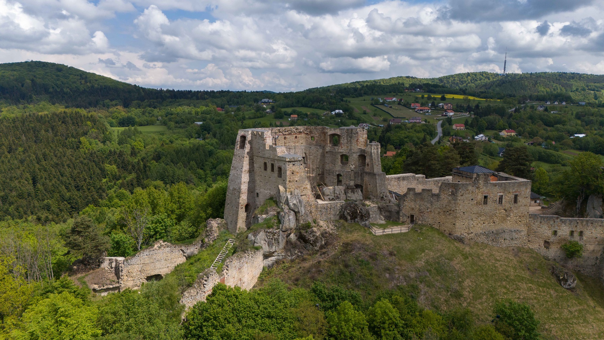 Panorama ruin zamku Ogrodzieniec z drewnianymi schodami, otoczonego zielenią wzgórz i lasów pod błękitnym niebem z obłokami.