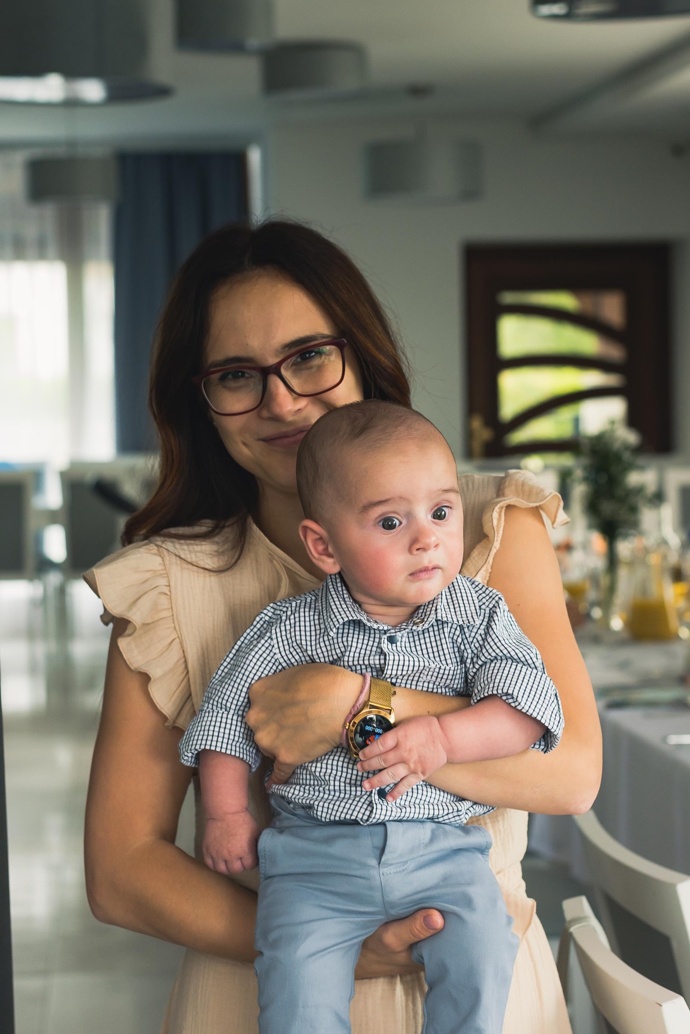 Kobieta w okularach trzyma na rękach niemowlę ubrane w koszulę w kratę i niebieskie spodnie. W tle widoczny stół z dekoracjami.