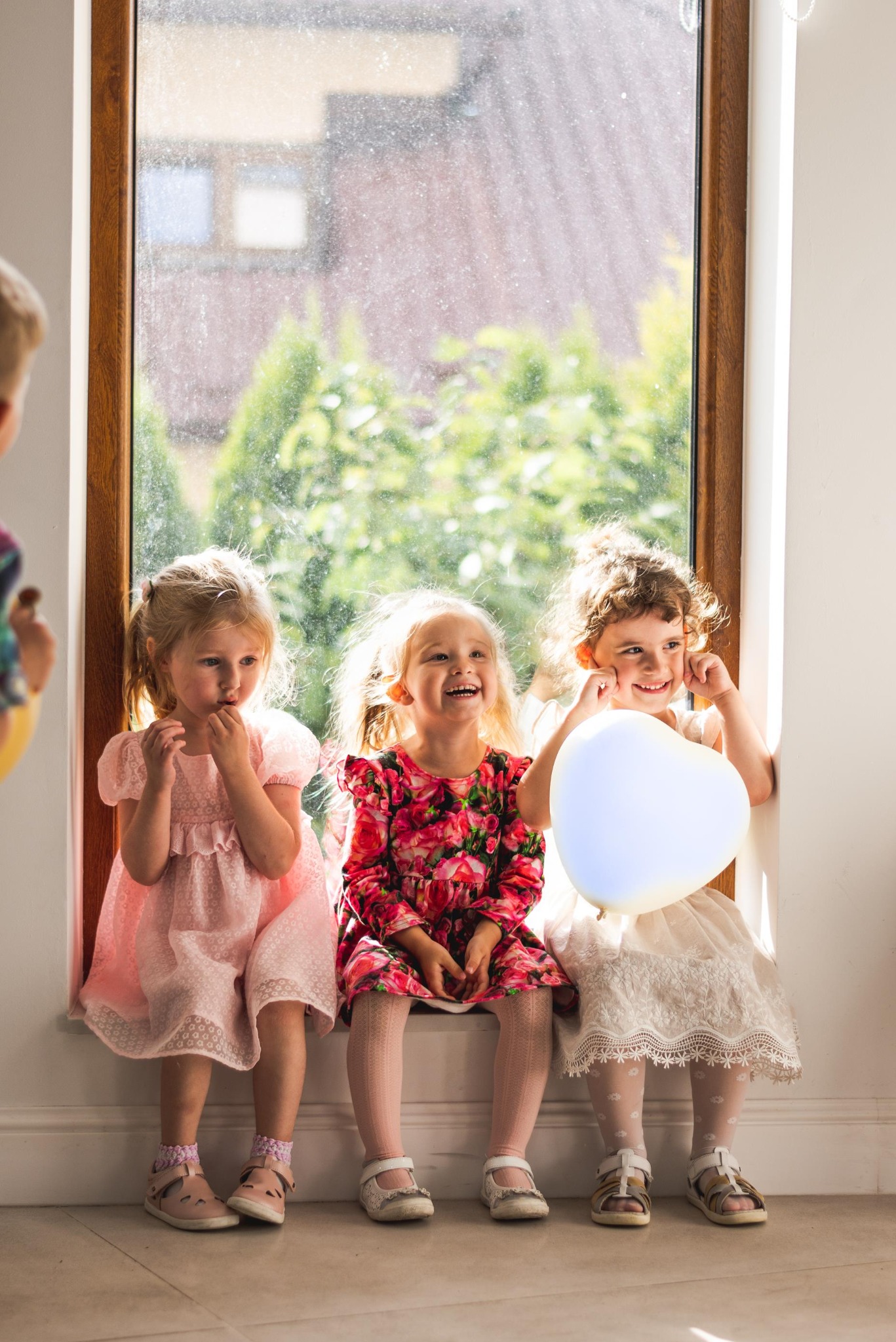 Trzy uśmiechnięte dziewczynki w sukienkach siedzą na parapecie okna. Jedna trzyma balon w kształcie serca. Jasne, naturalne światło. Fotografia dziecięca.