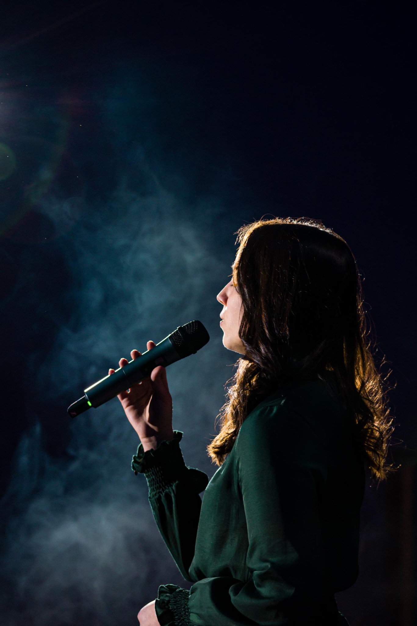 Kobieta śpiewa do mikrofonu na scenie w ciemności, z delikatnym oświetleniem z góry i efektem dymu w tle. Profil twarzy, zielona bluzka z marszczeniami.