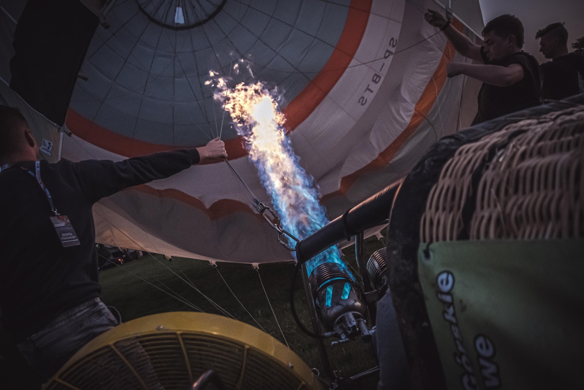 Napełnianie balonu na ogrzane powietrze: płomień palnika rozświetla wnętrze czaszy, widoczni ludzie obsługujący sprzęt. Ujęcie z dołu.