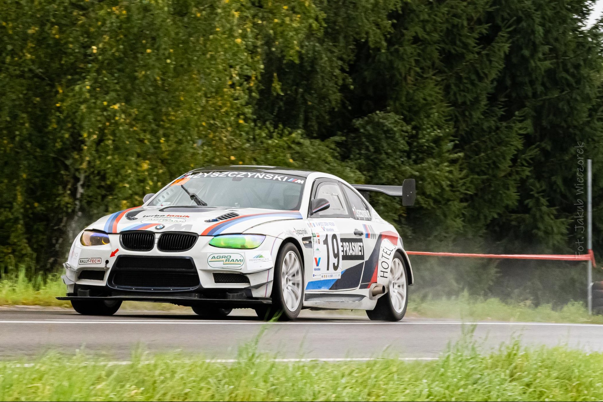 Białe BMW M3 w trakcie wyścigu, z numerem 19, na tle zieleni. Dynamiczne ujęcie z Krzeszowic. Widoczne logotypy sponsorów na karoserii.
