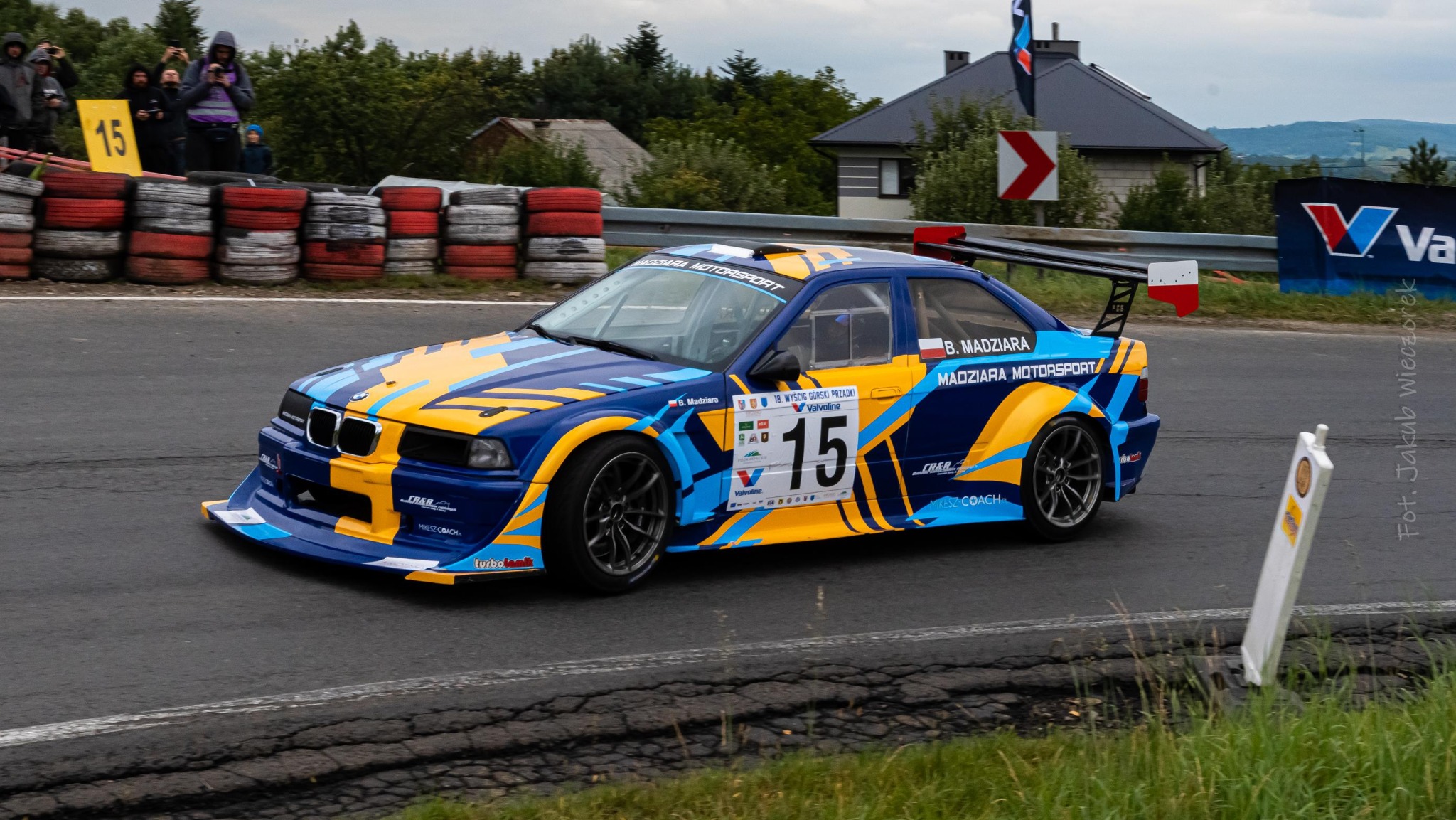 Dynamiczne ujęcie niebiesko-żółtego samochodu wyścigowego BMW E36 z numerem 15 na torze, z widocznymi oponami zabezpieczającymi i kibicami w tle. Ostry kadr, pochmurna pogoda.