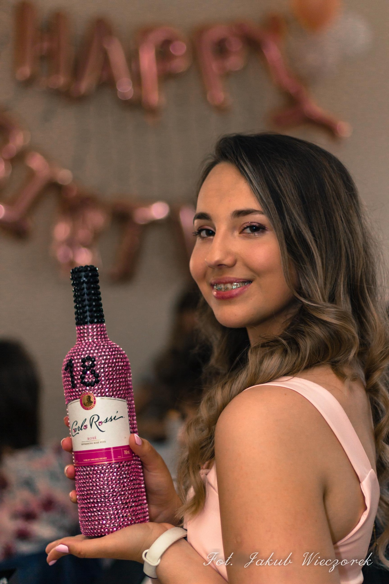 Uśmiechnięta osiemnastolatka z aparatem ortodontycznym trzyma błyszczącą, różową butelkę wina Carlo Rossi na tle dekoracji urodzinowych.