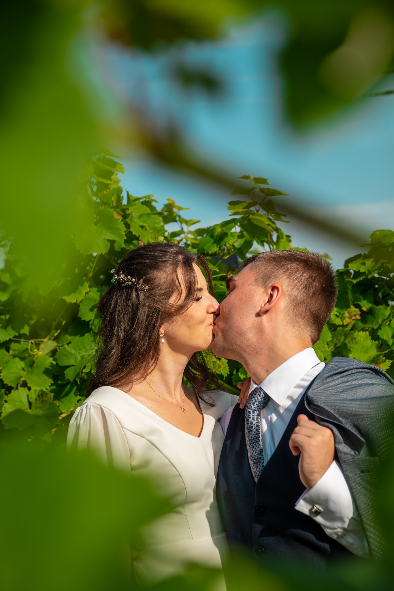 Para młoda w plenerze, pocałunek na tle zielonych liści. Panna młoda w białej sukni, pan młody w garniturze z marynarką na ramieniu. Romantyczna sesja.