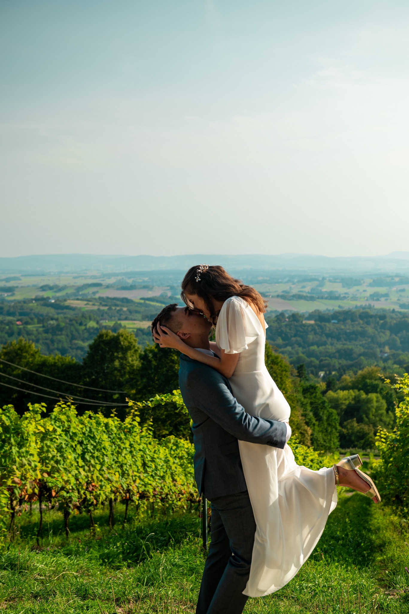 Para młoda w objęciach na tle winnicy i rozległego krajobrazu. Panna młoda w białej sukni, pan młody w szarym garniturze. Ujęcie z dołu, podkreślające radość chwili.
