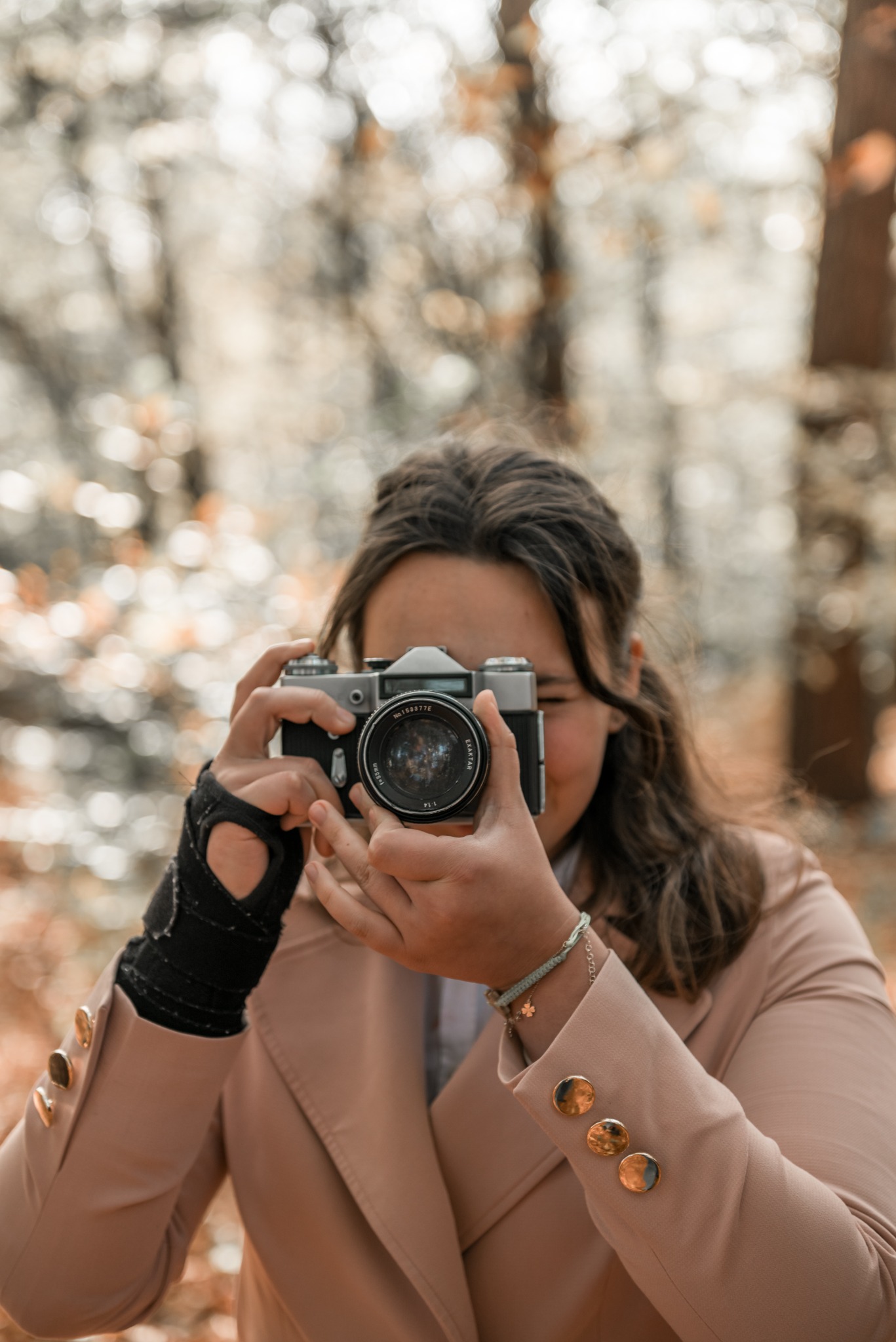 Kobieta w beżowej marynarce z mankietem na ręce robi zdjęcie aparatem analogowym na tle rozmytego, jesiennego lasu.