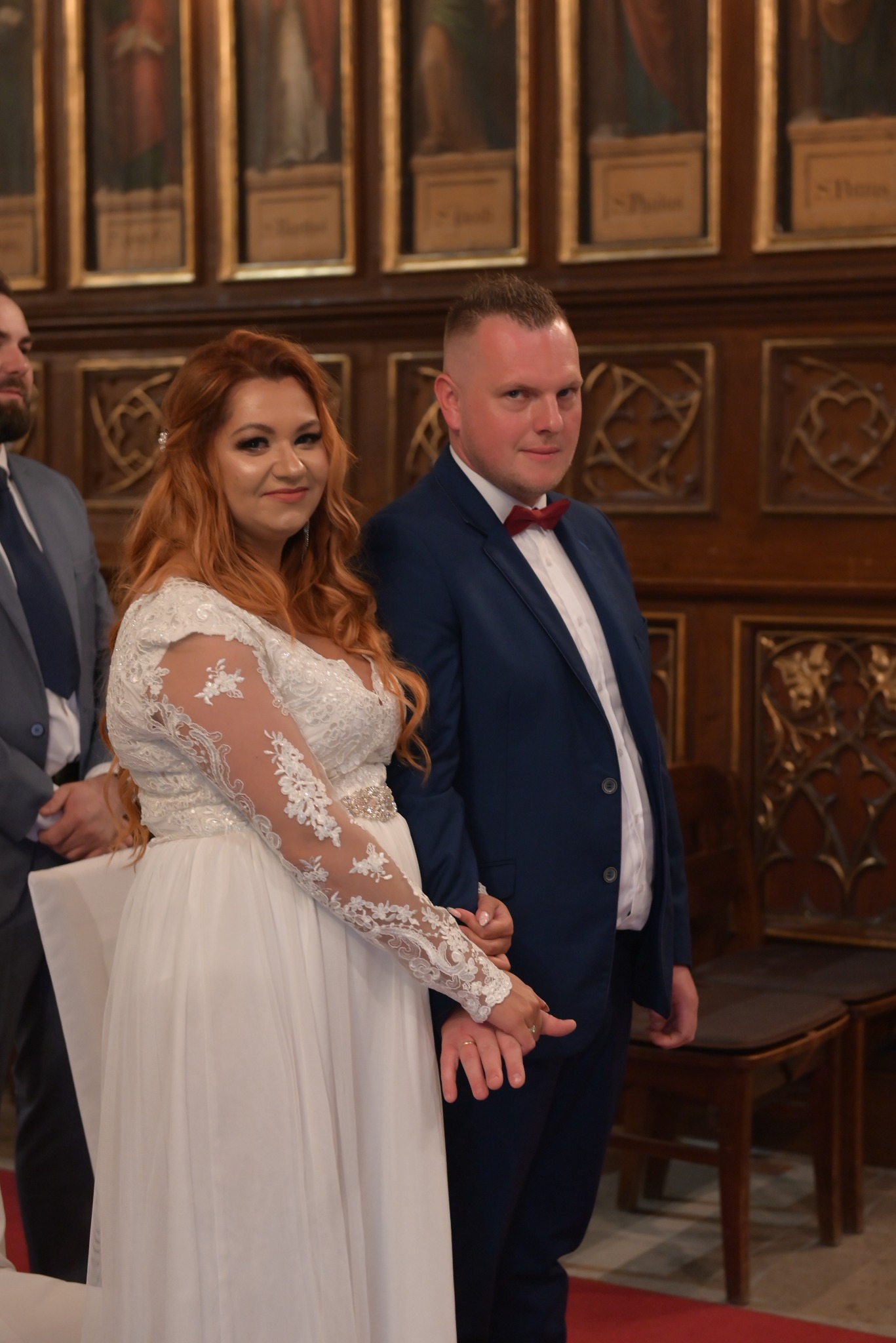Para młoda w kościele: panna młoda w koronkowej sukni z długim rękawem, pan młody w granatowym garniturze i bordowej muszce. W tle drewniane dekoracje i obrazy.