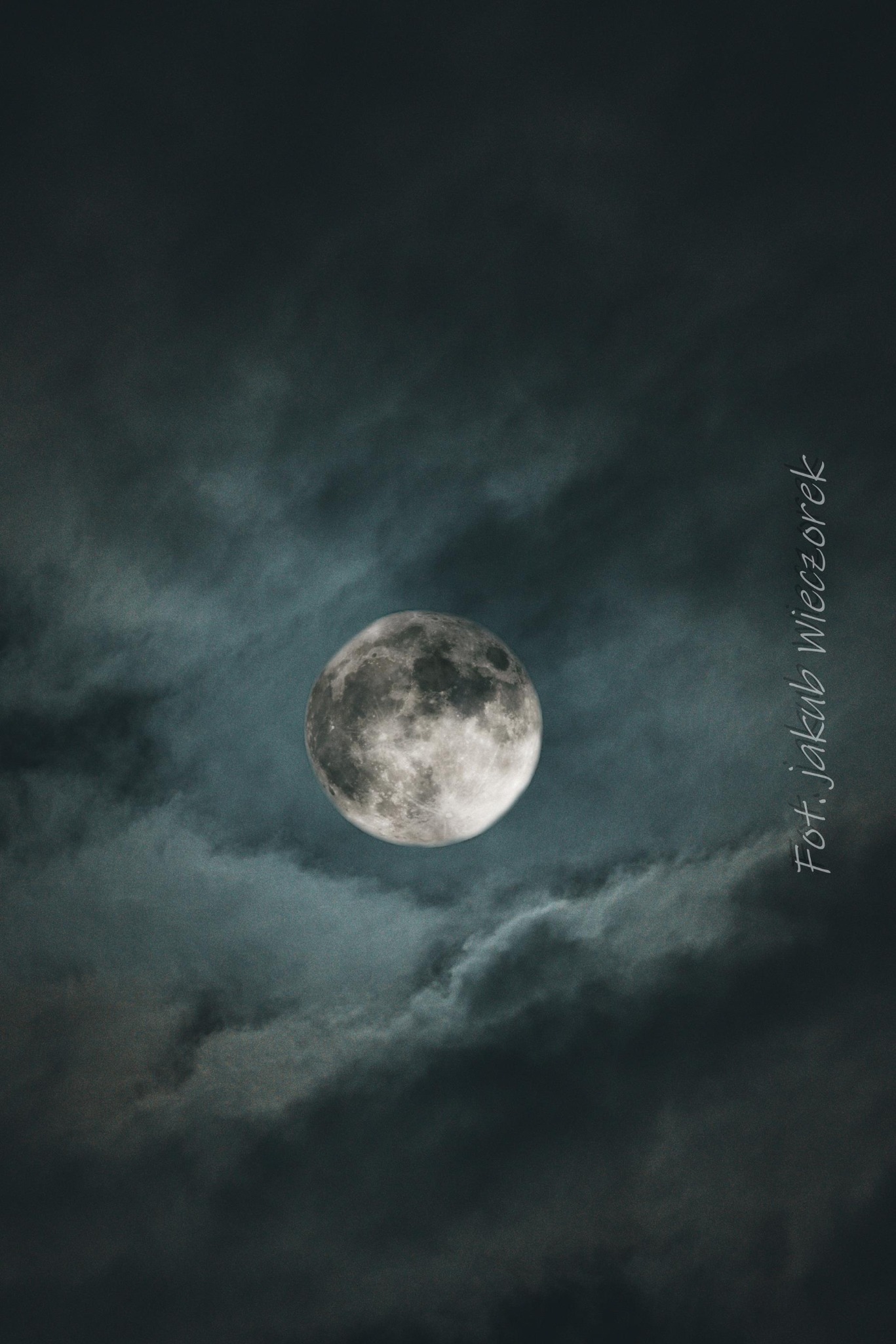 Księżyc w pełni, otoczony ciemnymi chmurami, sfotografowany z dołu. W prawym dolnym rogu widoczny podpis autora zdjęcia: Fot. Jakub Wieczorek.
