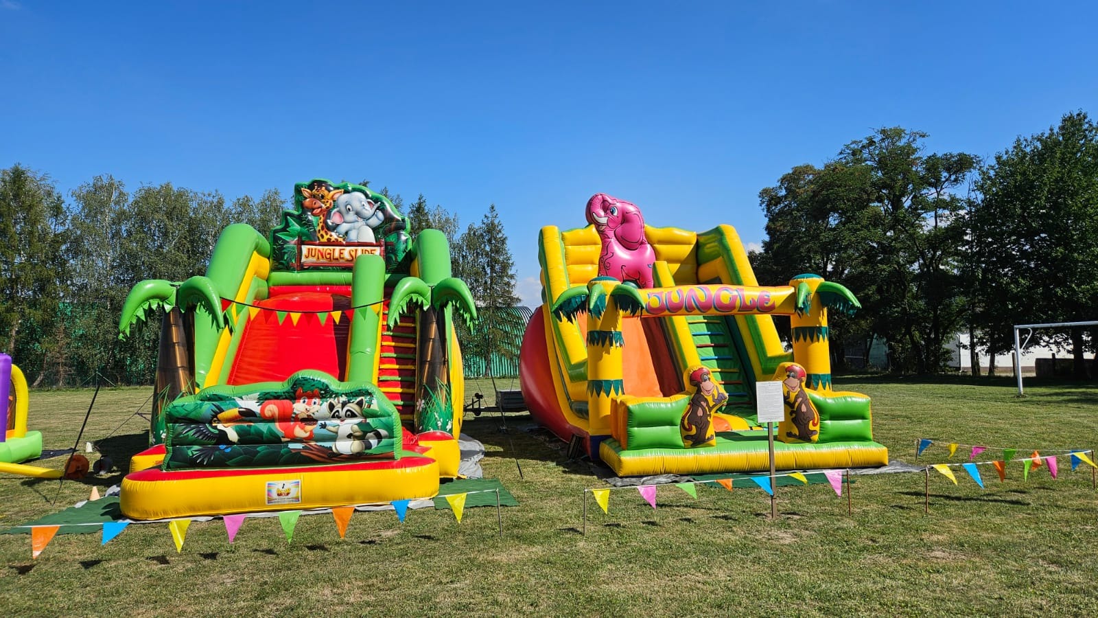 Dwa dmuchane place zabaw 'Jungle Slide' na trawie, z motywami zwierząt dżungli i kolorowymi girlandami, pod błękitnym niebem. Park rozrywki.