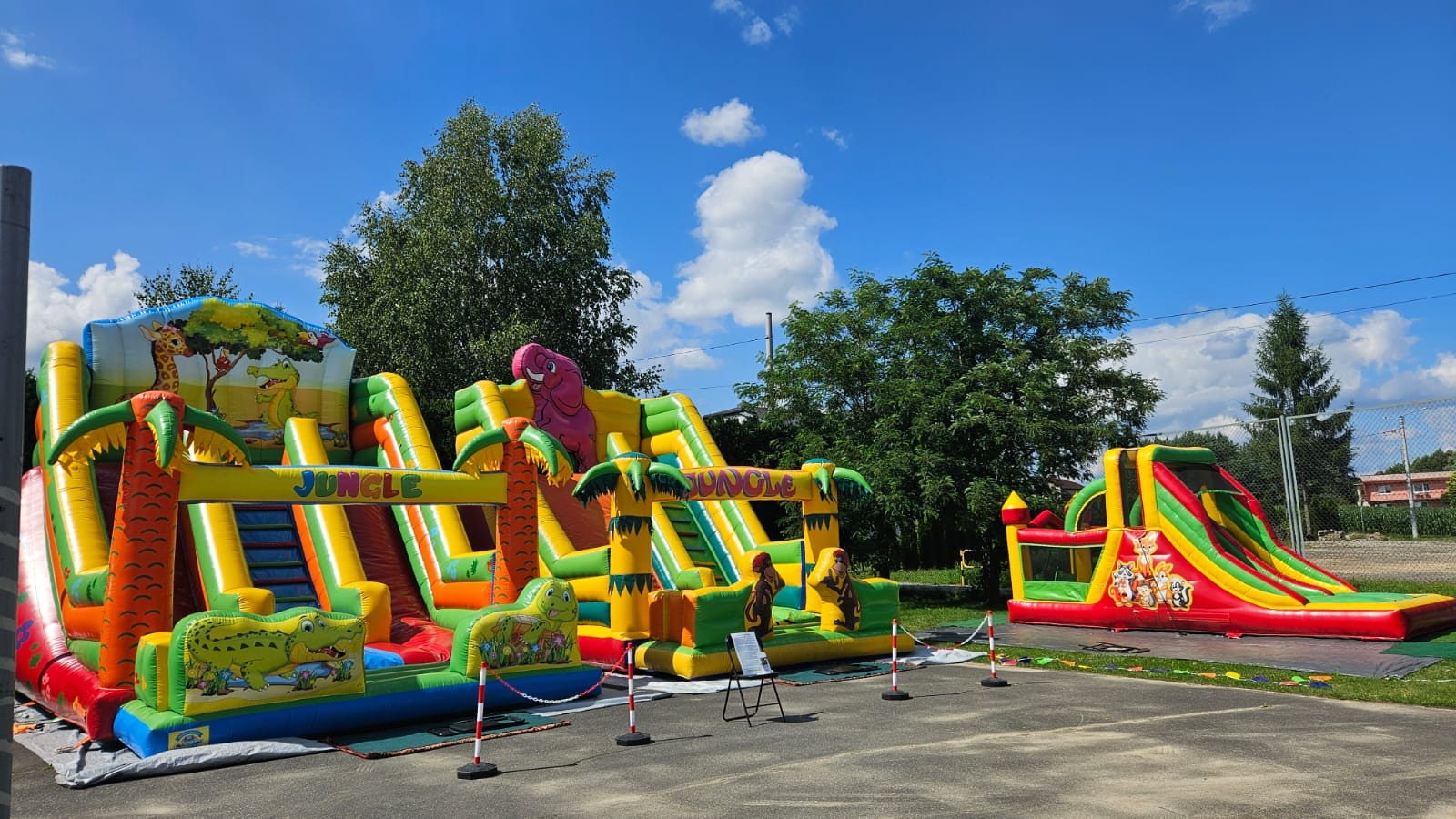 Kolorowe dmuchańce z motywem dżungli: zjeżdżalnie, palmy, krokodyle i słoń. Atrakcje na imprezę plenerową, widoczne barierki.