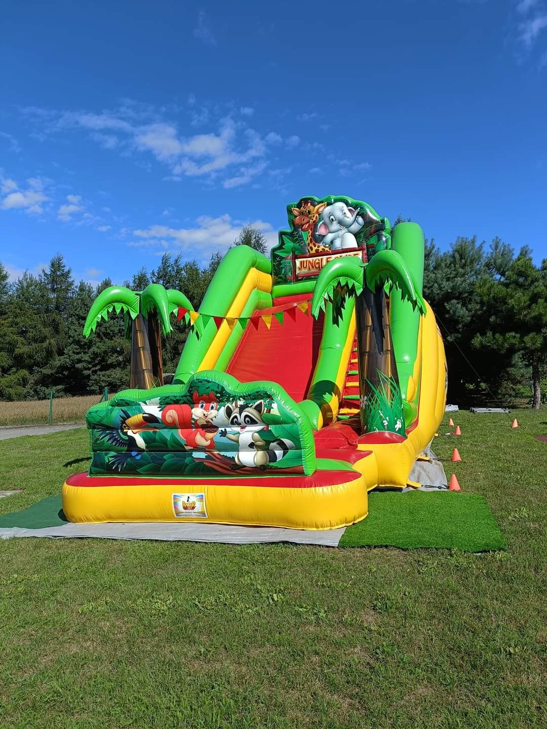 Kolorowa, dmuchana zjeżdżalnia w stylu dżungli na trawie, z motywami zwierząt i palm, na tle błękitnego nieba. Widoczne pomarańczowe pachołki.