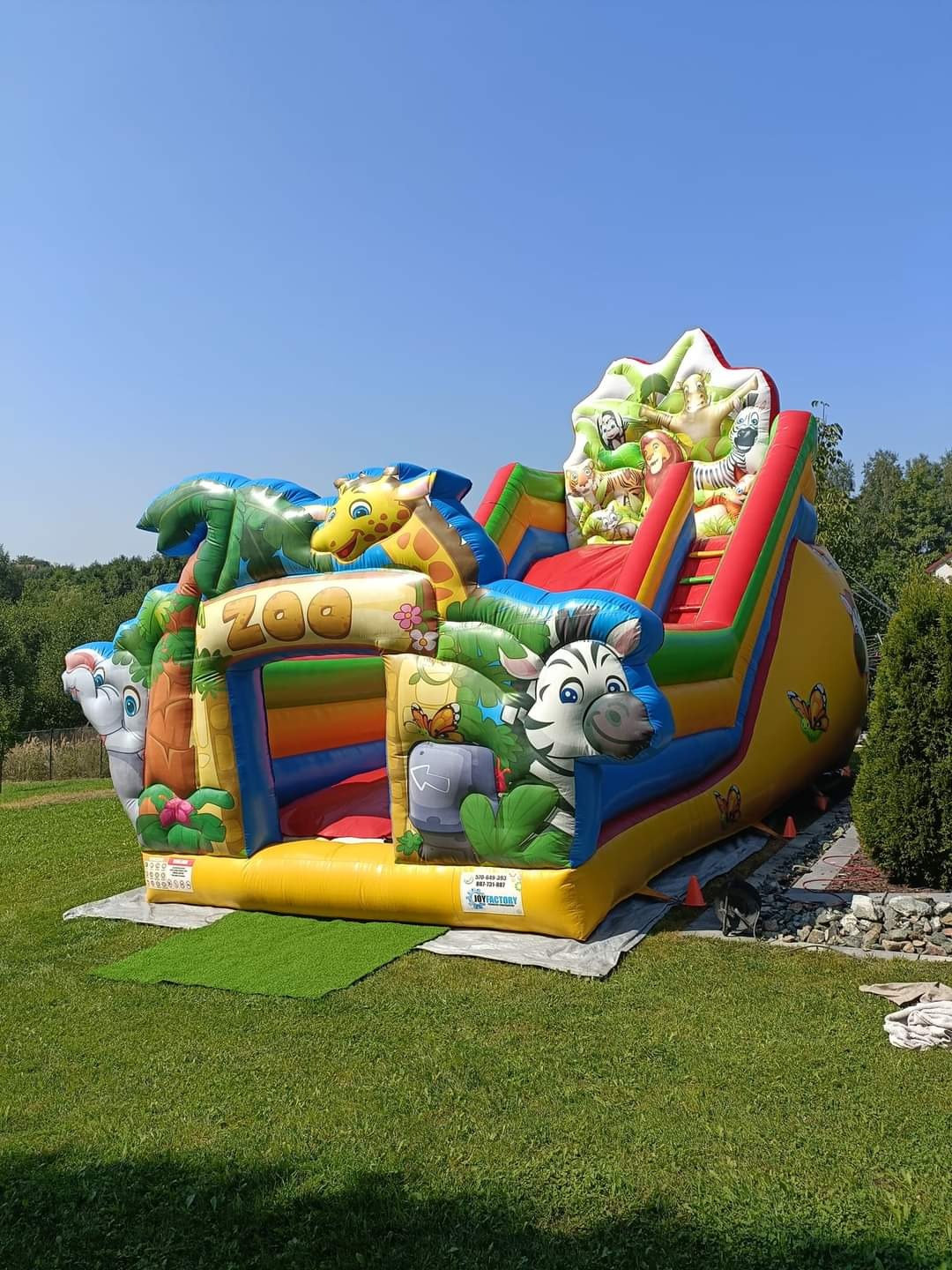 Kolorowy, duży dmuchany zamek ze zjeżdżalnią z motywem zoo na trawie. Widoczne postacie zwierząt: żyrafa, zebra, lew, małpa. Słoneczny dzień, błękitne niebo w tle.