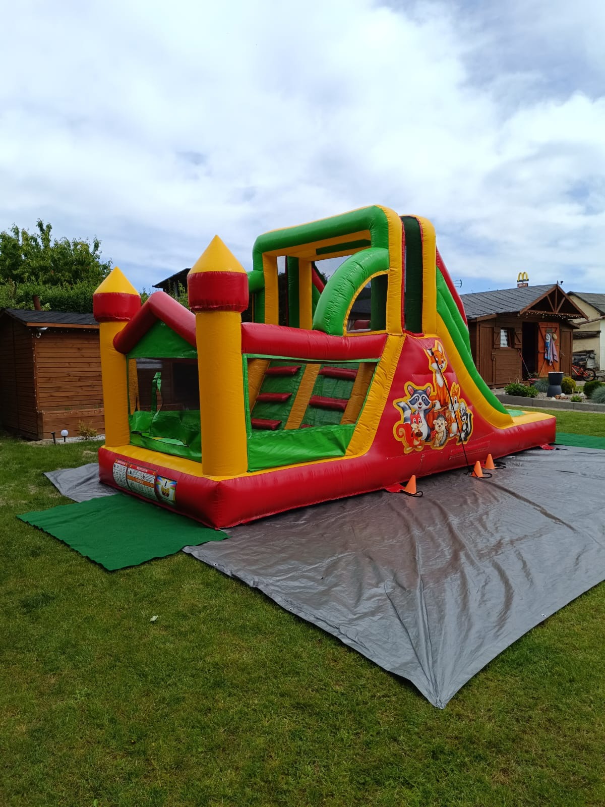 Kolorowy, rozłożony na trawie dmuchany zamek ze zjeżdżalnią, z motywem leśnych zwierząt, zabezpieczony pomarańczowymi pachołkami. Widok z niskiej perspektywy.