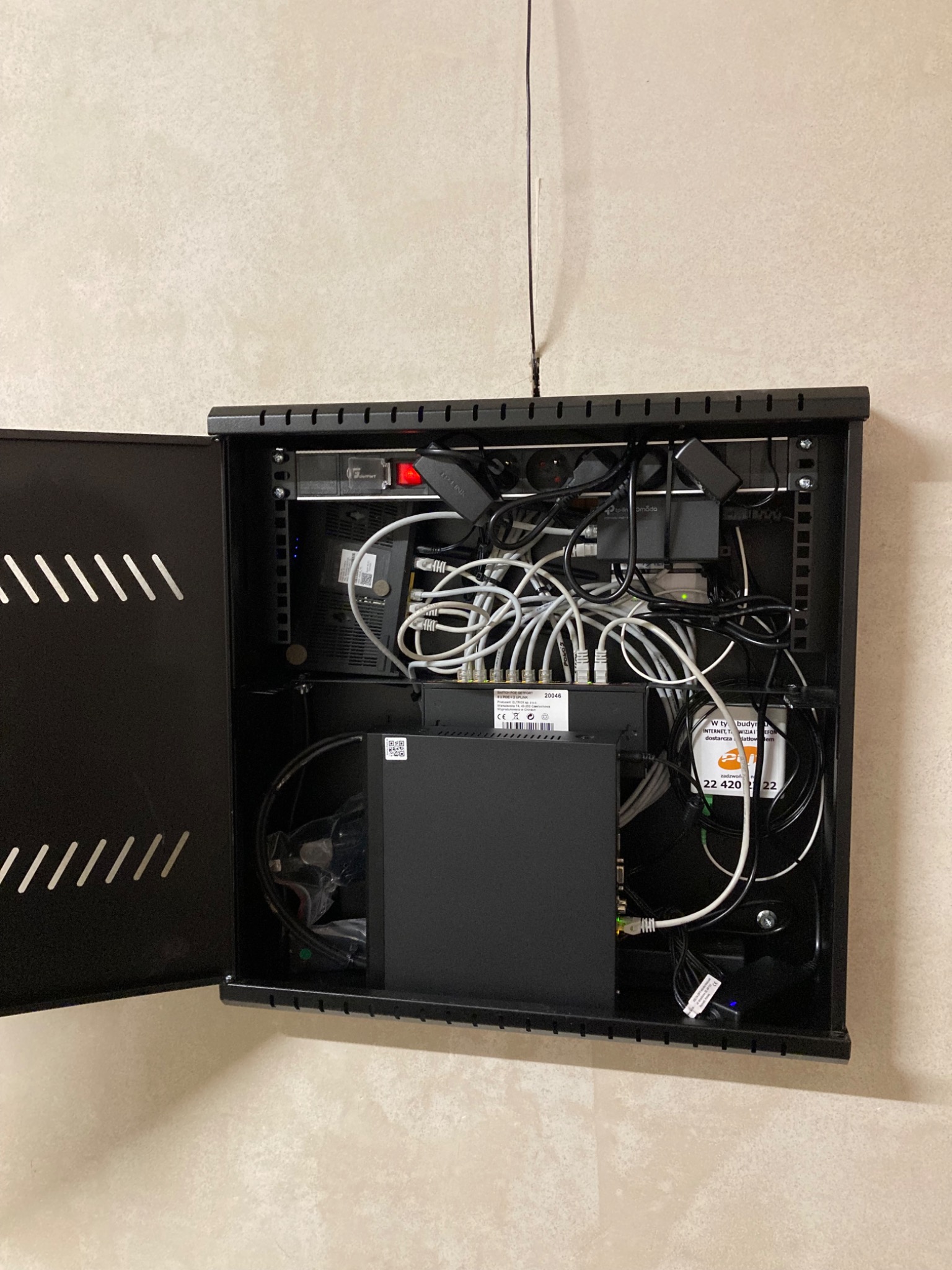 Otwarta, czarna szafka rack na ścianie z licznymi kablami i urządzeniami sieciowymi wewnątrz. Widoczne zasilacze, routery i przewody. Antena wystaje z górnej części szafki.