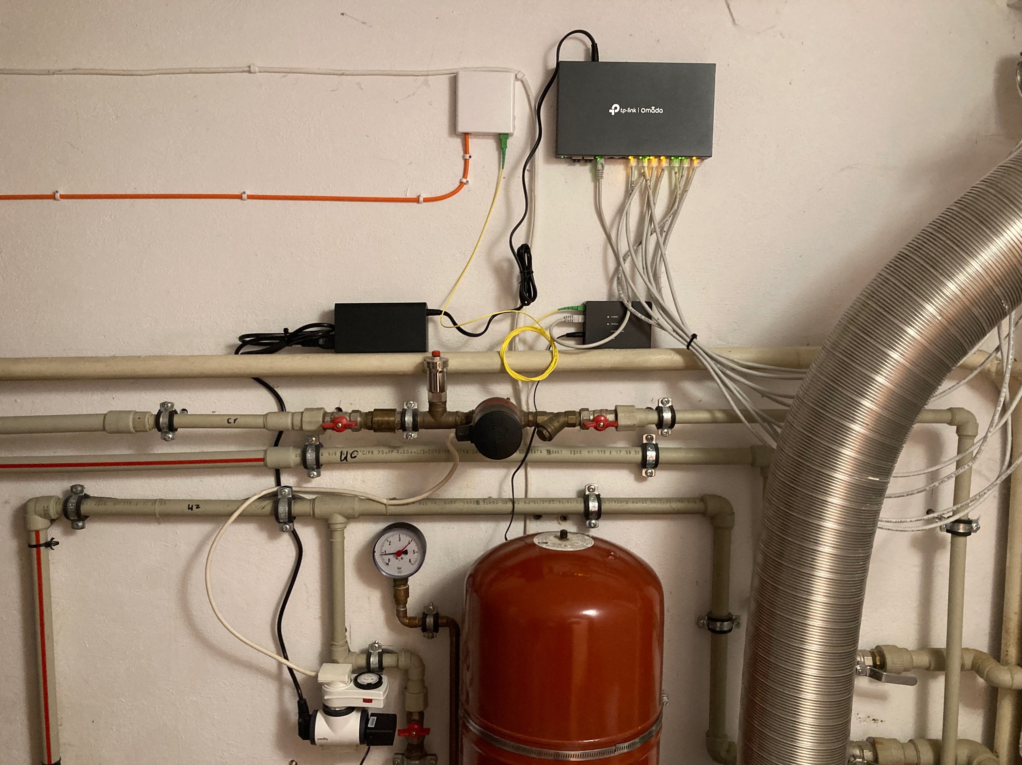 Rozdzielnica telekomunikacyjna TP-Link Omada z podłączonymi kablami Ethernet, obok rury instalacyjne i przewody, na białej ścianie.