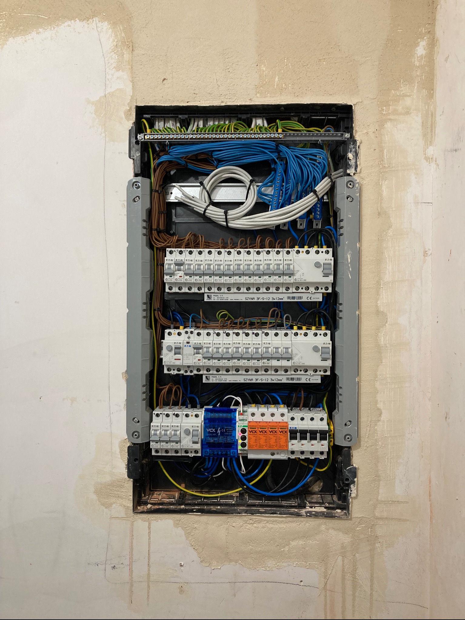 Rozdzielnica elektryczna w trakcie montażu w ścianie, widoczne kable i zabezpieczenia. Ściana wokół rozdzielnicy jest w stanie surowym, przygotowana do wykończenia.