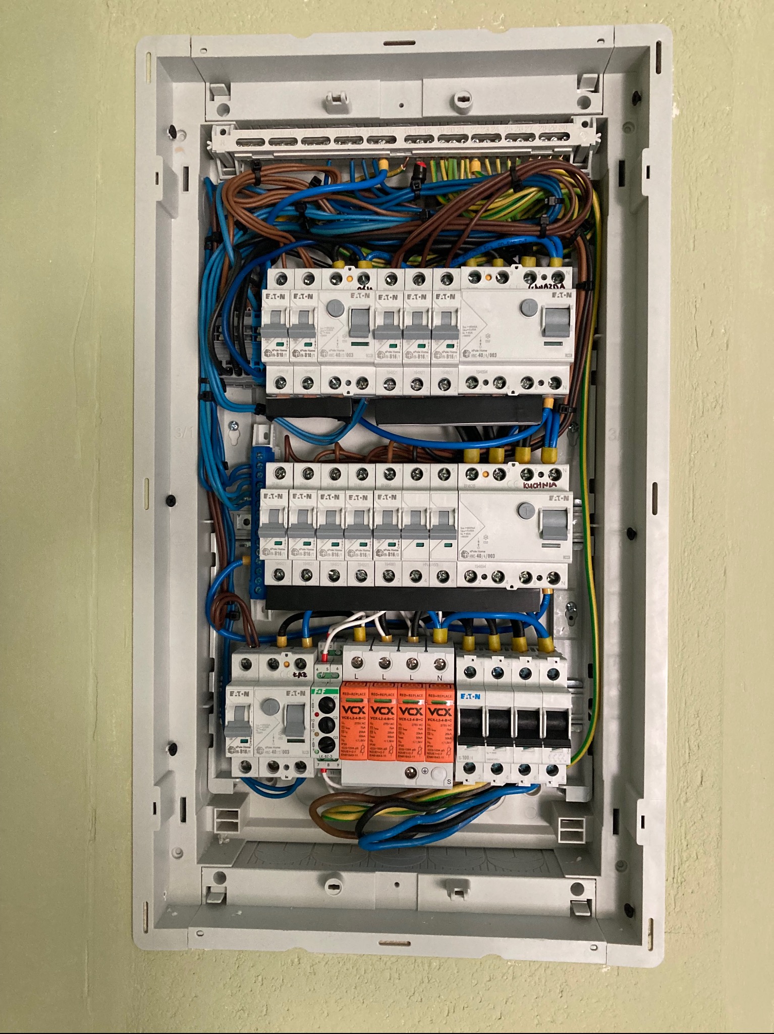 Rozdzielnica elektryczna w obudowie, z widocznymi bezpiecznikami, przewodami w kolorach brązowym, niebieskim i żółto-zielonym. Widoczne elementy marki Eaton i Vox.