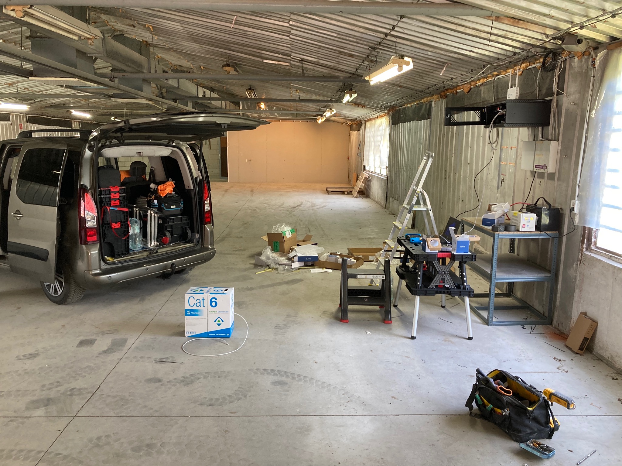 Instalacja kabli CAT6 w garażu: otwarty bagażnik auta z narzędziami, drabina, stół roboczy z akcesoriami, skrzynka kabli. Widoczne okablowanie przy ścianie.