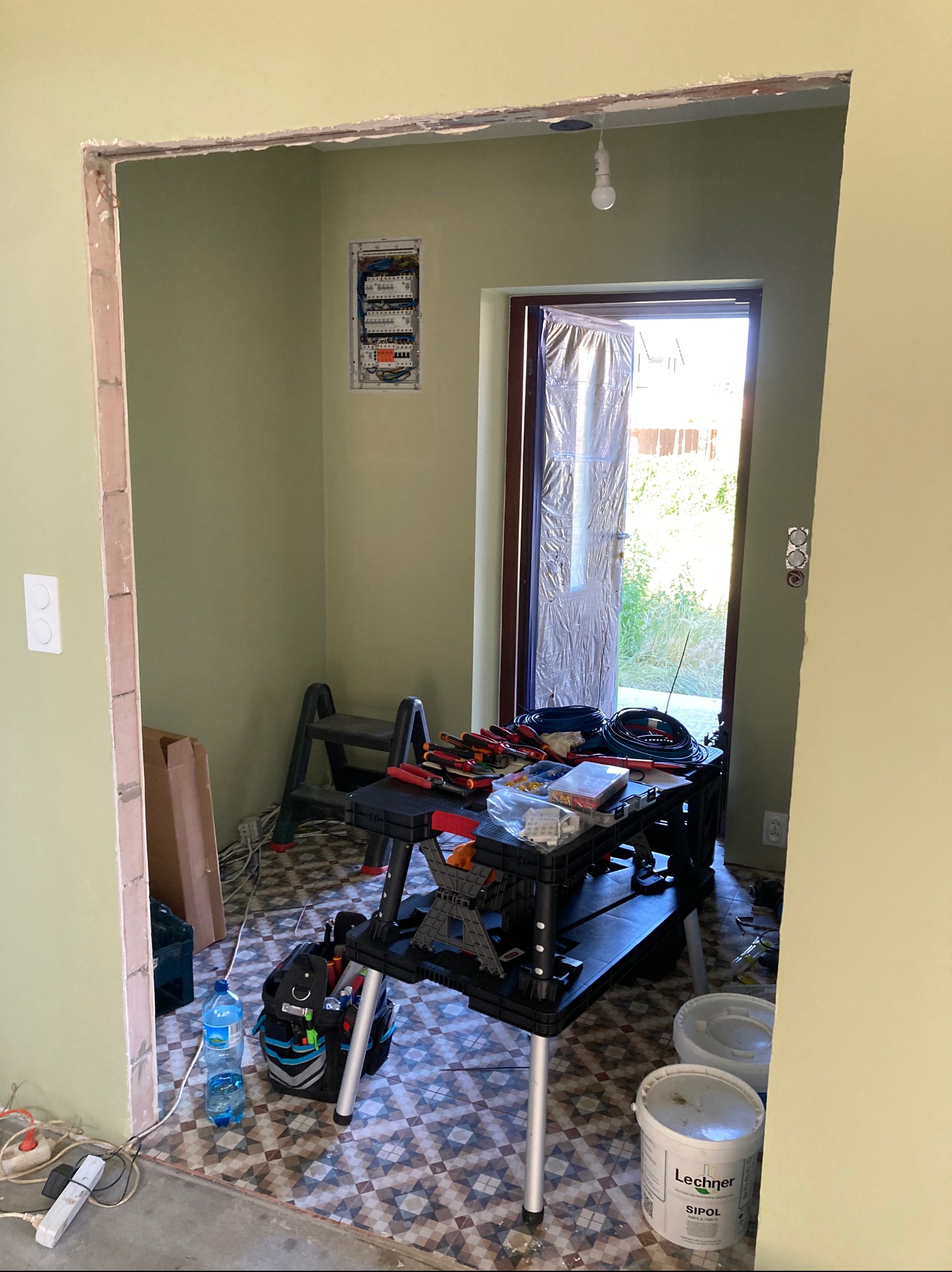 Wnętrze w trakcie prac elektrycznych: rozdzielnia na ścianie, rozłożone narzędzia na stole roboczym, widoczna drabina i kable. Przygotowanie do instalacji elektrycznej.