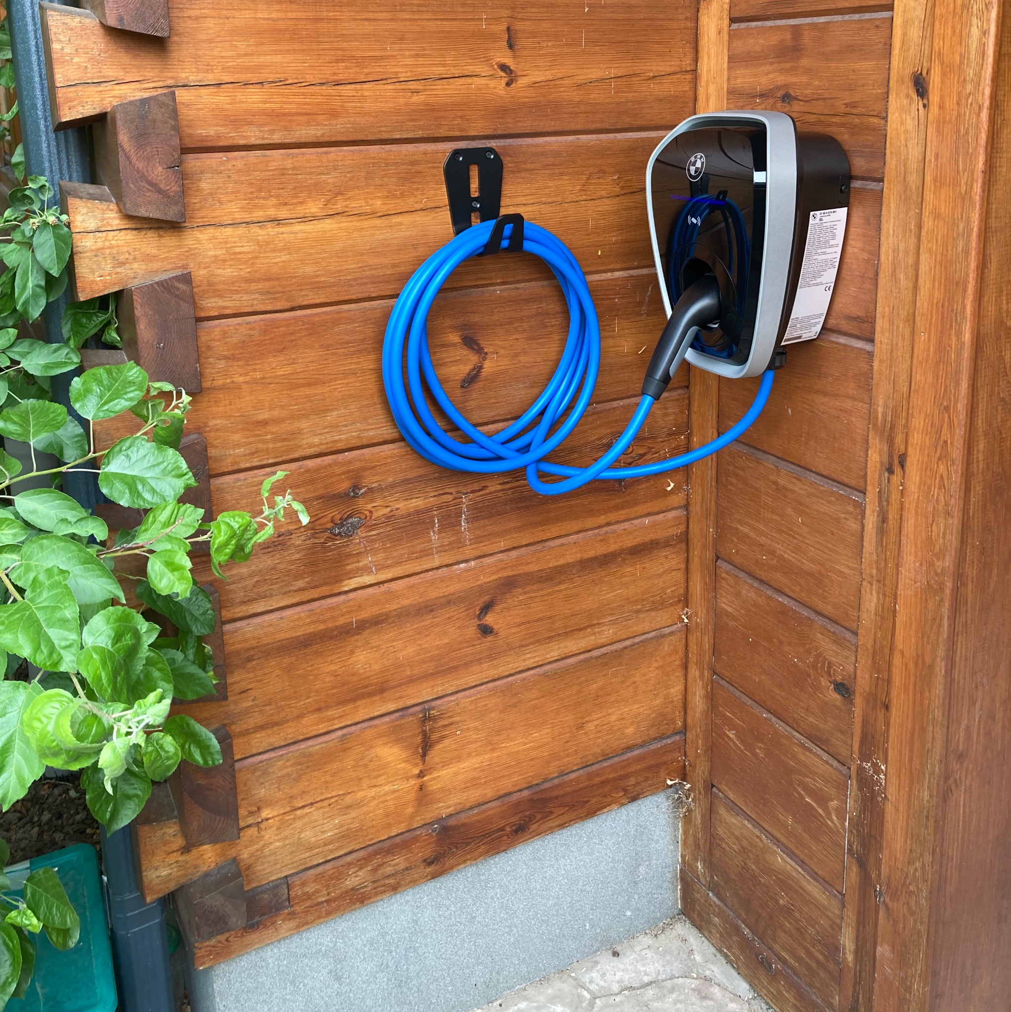 Na drewnianej elewacji zamontowana stacja ładowania pojazdów elektrycznych BMW z niebieskim kablem. Przyłącze na czarnym uchwycie. Obok roślinność.
