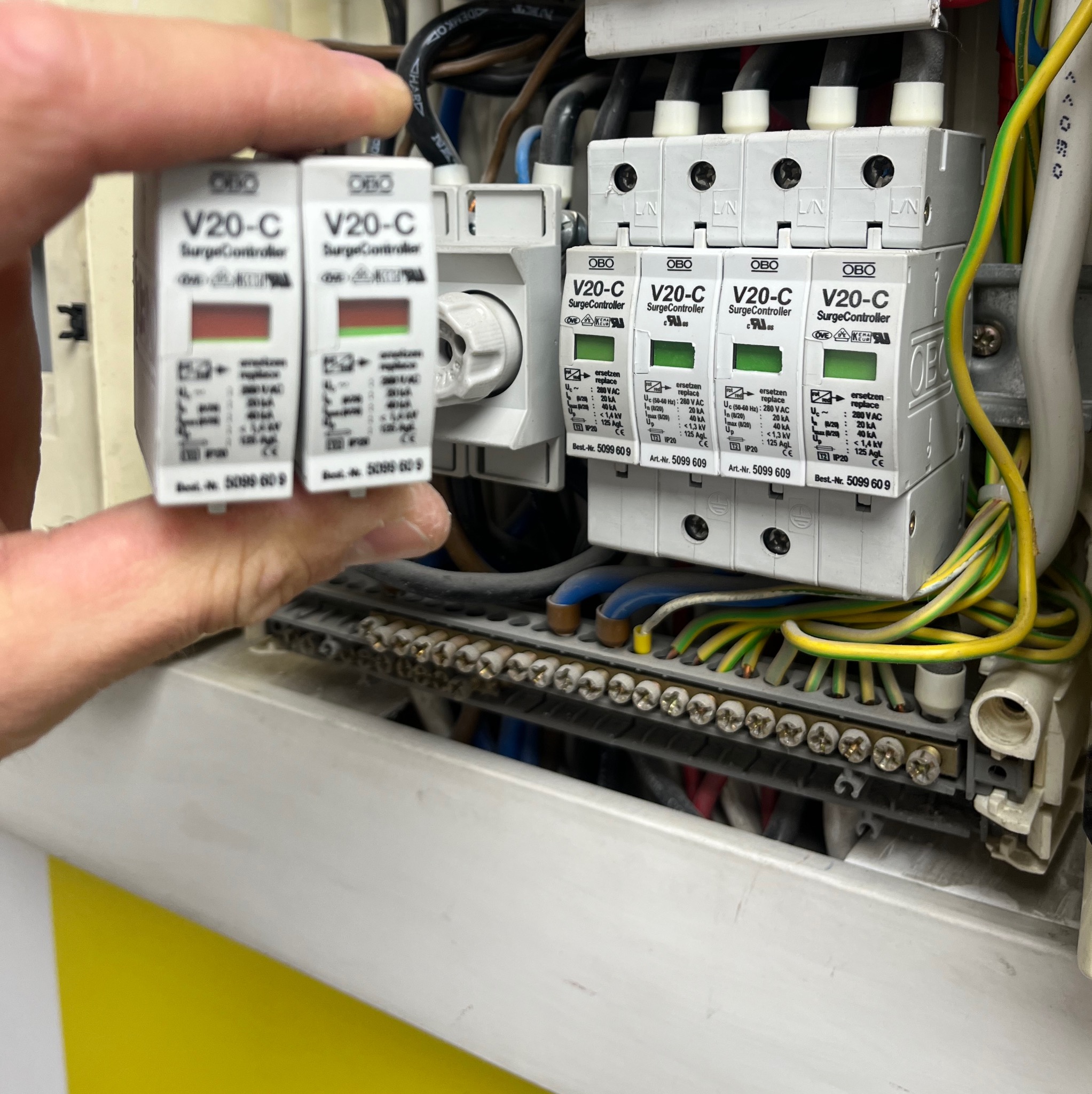 Instalacja zabezpieczeń przeciwprzepięciowych OBO V20-C w rozdzielnicy elektrycznej. Widoczne przewody i listwa zaciskowa. Dwa kontrolery trzymane w dłoni.