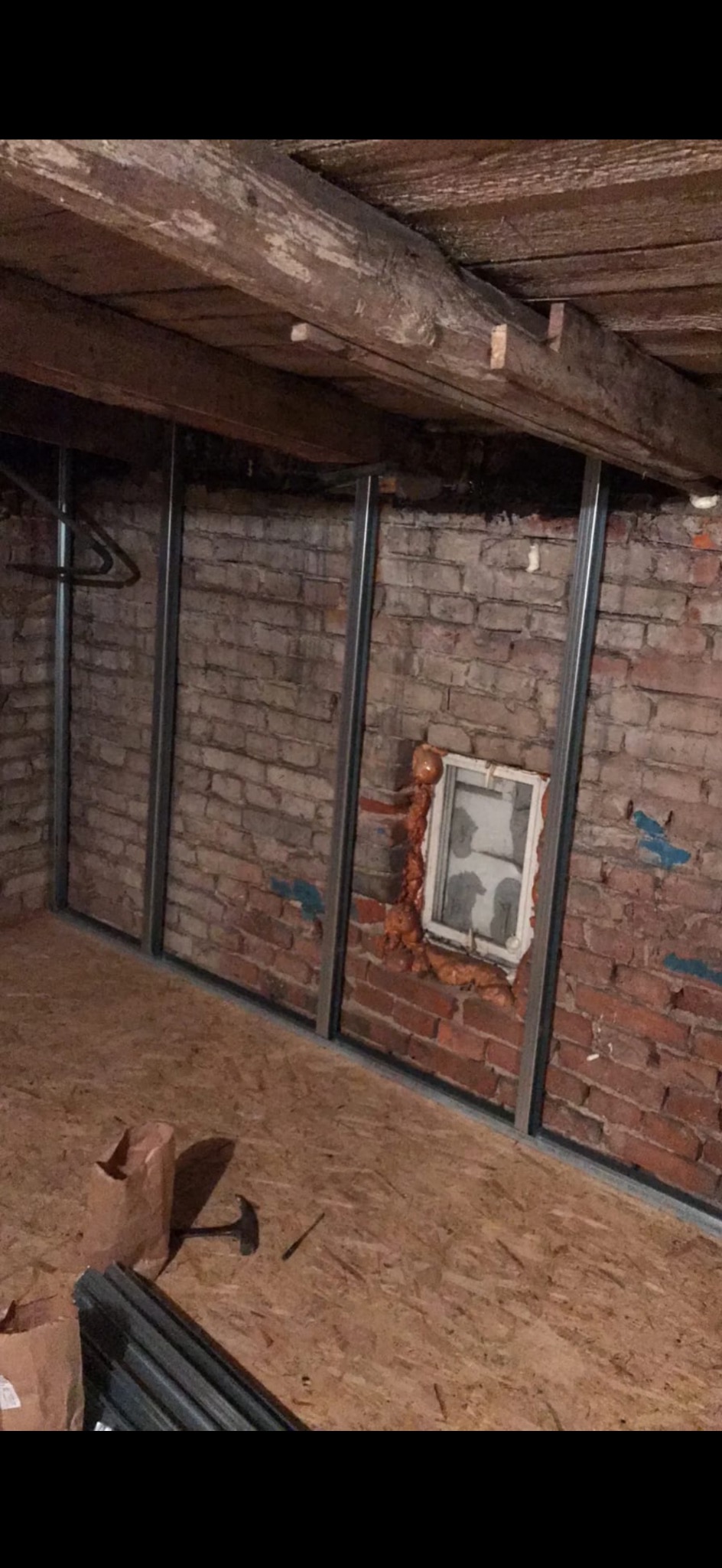 Ceglana ściana z oknem uszczelnionym pianką, na tle surowego drewnianego stropu. Widoczne metalowe profile konstrukcyjne i fragment podłogi z płyty OSB.