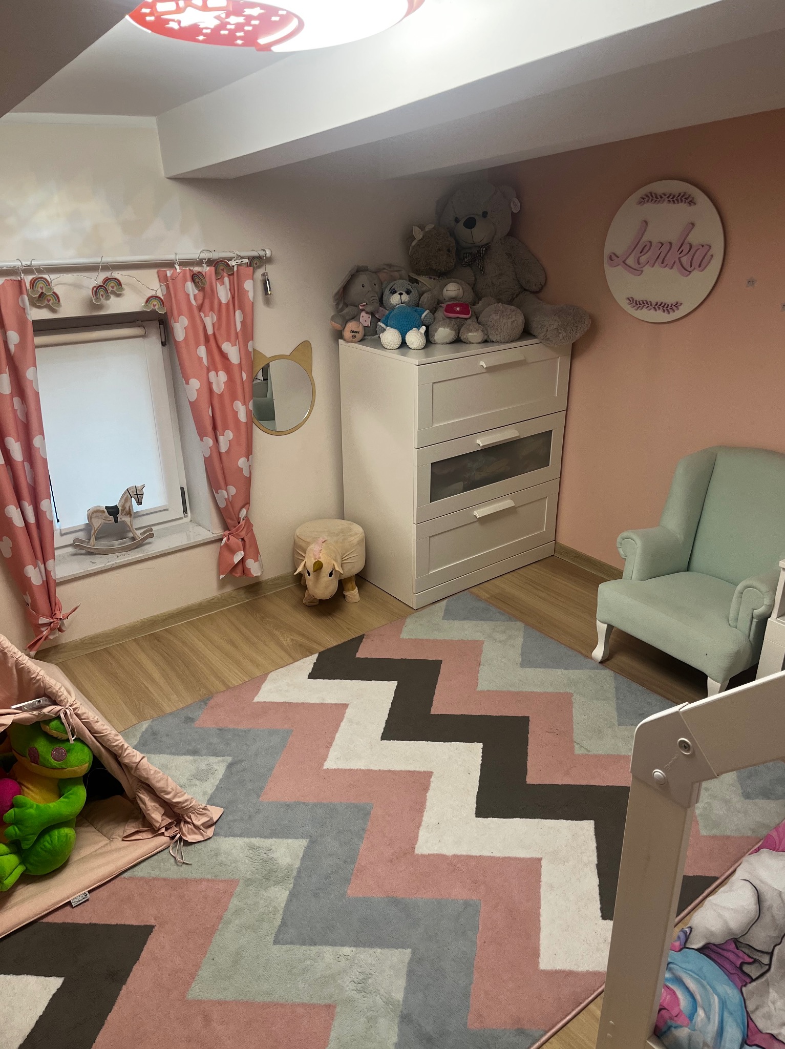 Pokój dziecięcy w pastelowych barwach: łóżko, komoda z pluszakami, fotel i dywan w zygzaki. Dekoracje okienne i lampa sufitowa dodają uroku.