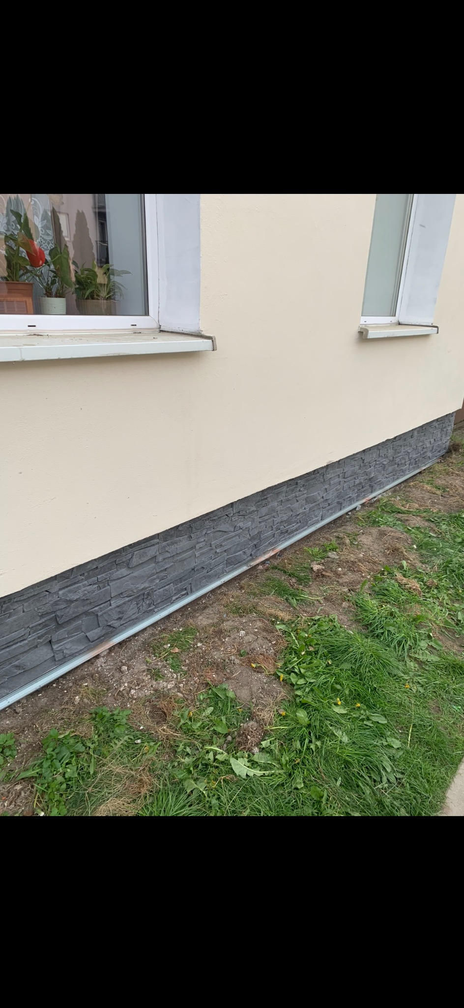 Fragment elewacji domu z oknami, wykończony szarym, kamiennym cokołem. Widoczne rośliny w oknie i trawa przy ścianie. Ujęcie z poziomu gruntu.