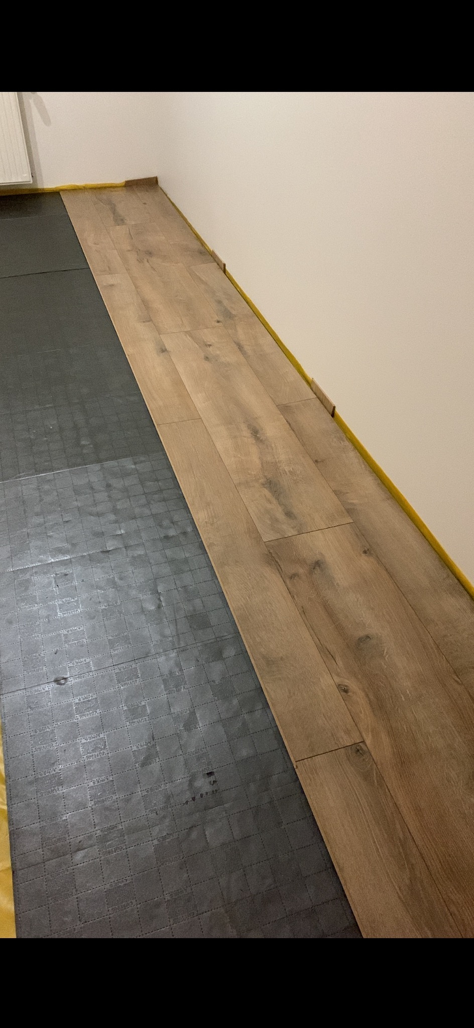 Układanie paneli podłogowych wzdłuż ściany zabezpieczonej żółtą taśmą, widoczna podkładka pod panele. Parkiet w odcieniach brązu i beżu.