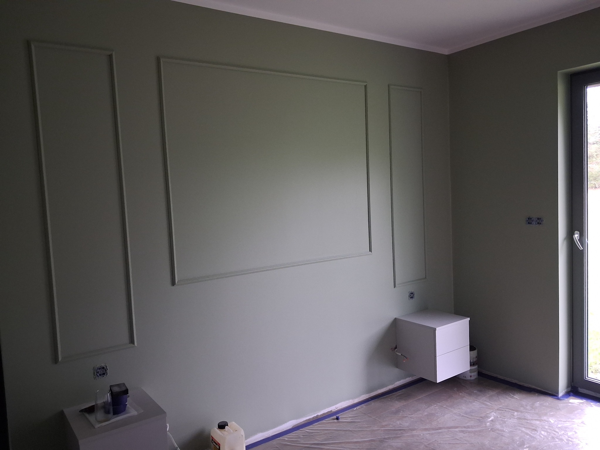 Ściana w odcieniach zieleni z dekoracyjnymi listwami i pomalowanymi gniazdkami, zabezpieczona folią malarską. Gotowe wykończenie wnętrza.