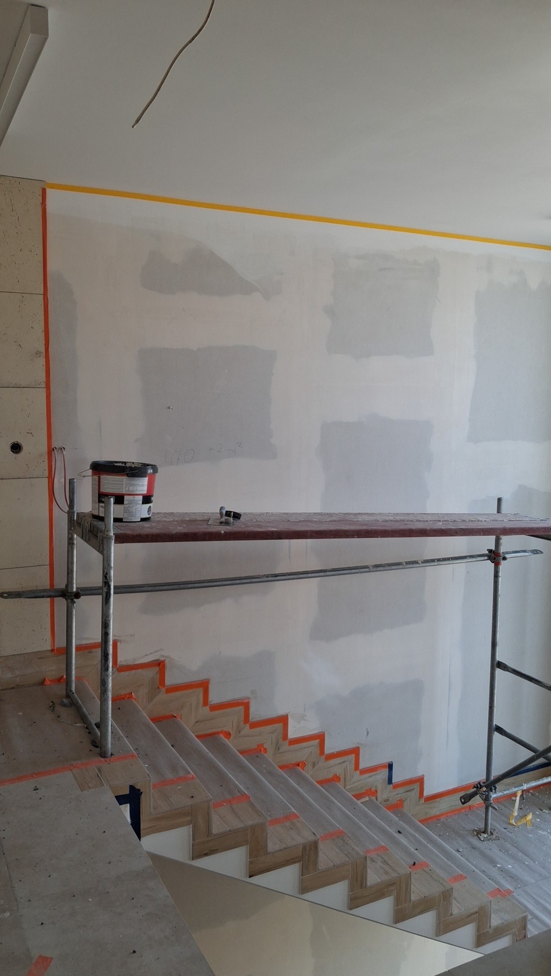 Ściana po szpachlowaniu, zabezpieczone taśmą schody i listwa, rusztowanie z farbą. Etap prac wykończeniowych w pomieszczeniu ze schodami.