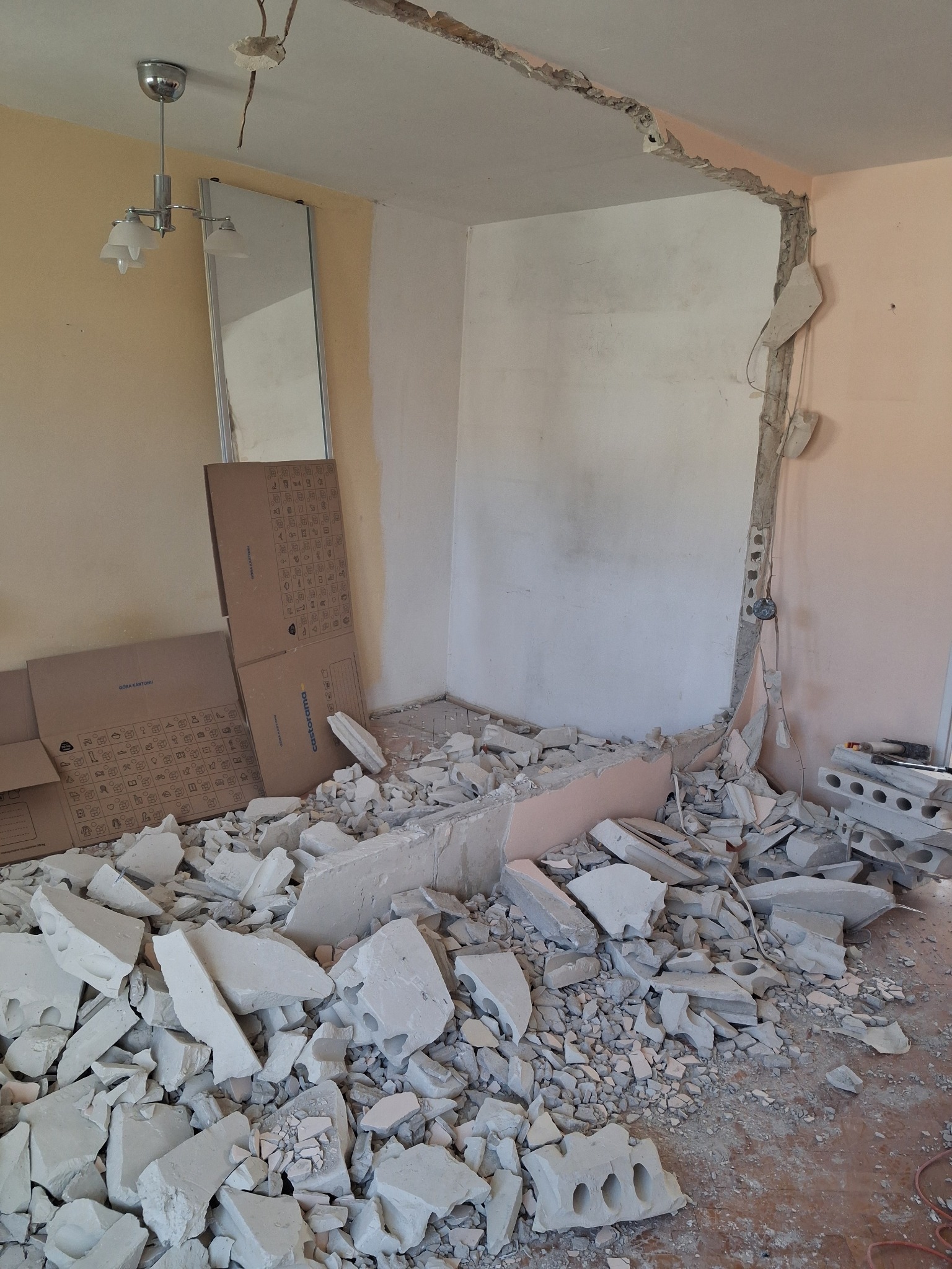 Fragment zdemontowanej ściany z widocznymi przewodami i gruzem na podłodze w pomieszczeniu z lampą i lustrem. Etap prac wyburzeniowych.