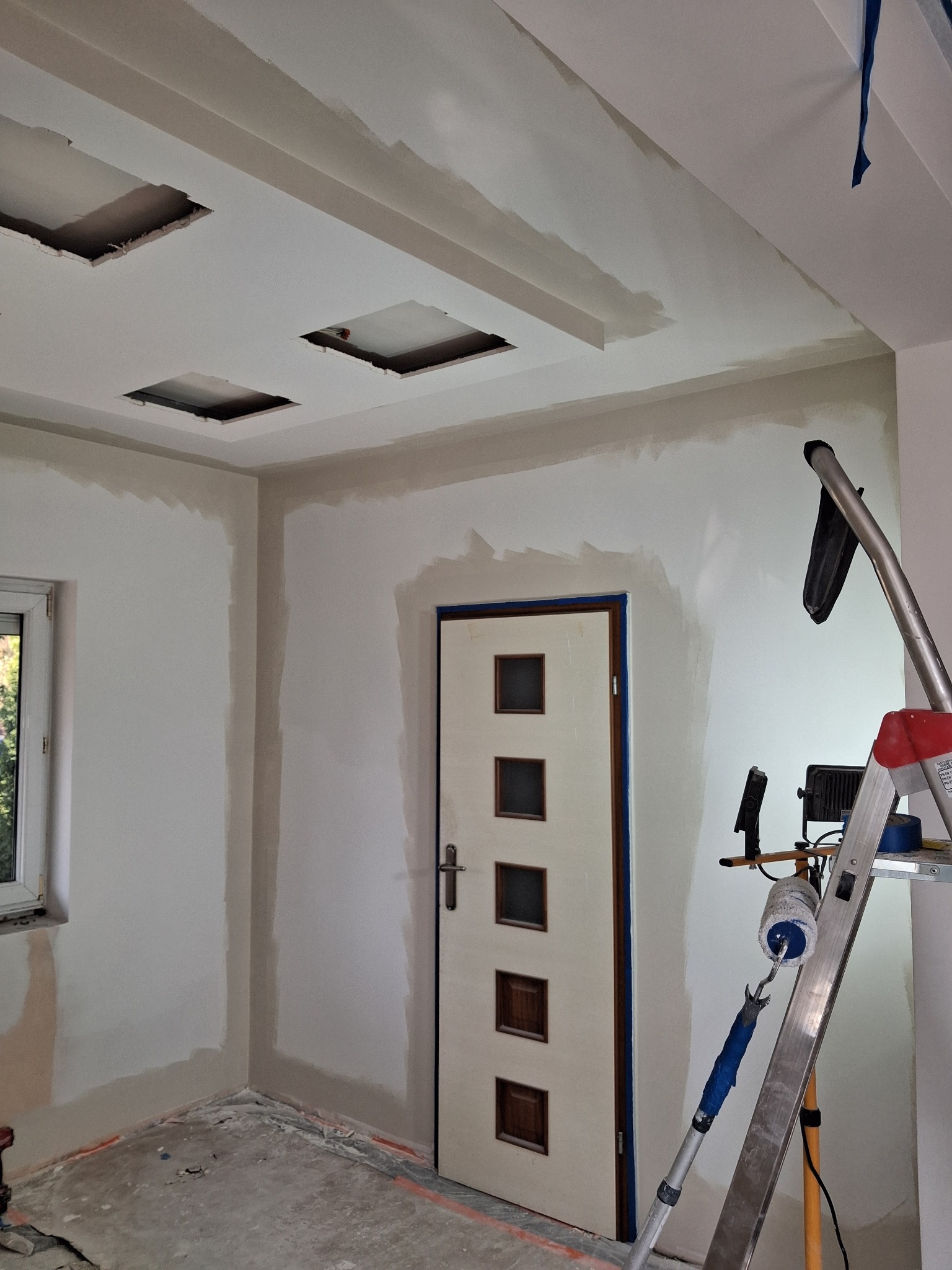 Pokój w trakcie prac przygotowawczych do malowania: ściany i sufit po szpachlowaniu, widoczna drabina malarska z wałkiem oraz taśma malarska na futrynie drzwi.