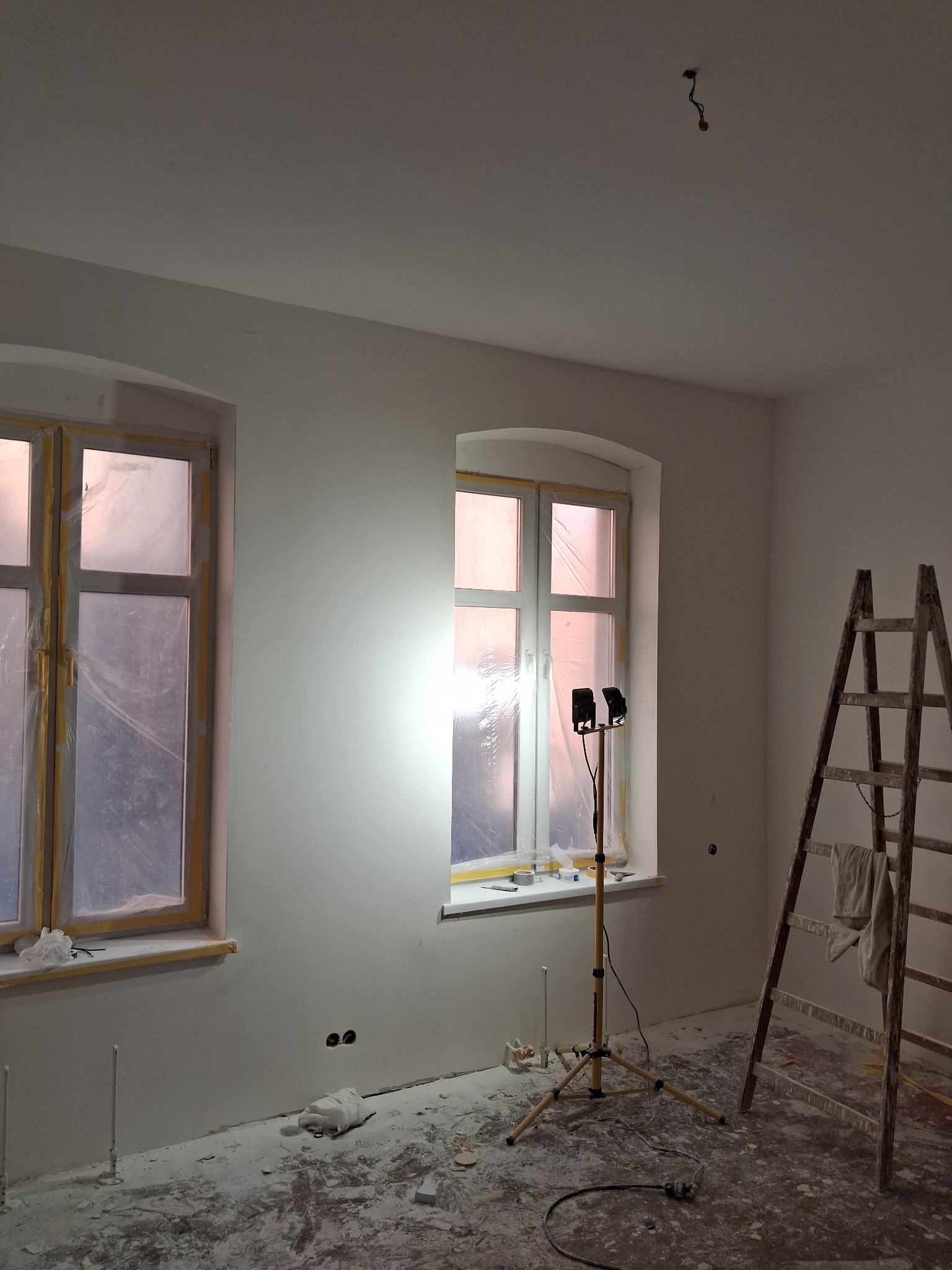 Pokój po szpachlowaniu ścian, okna zabezpieczone folią, widoczna drabina i lampa oświetleniowa. Na podłodze resztki materiałów budowlanych.