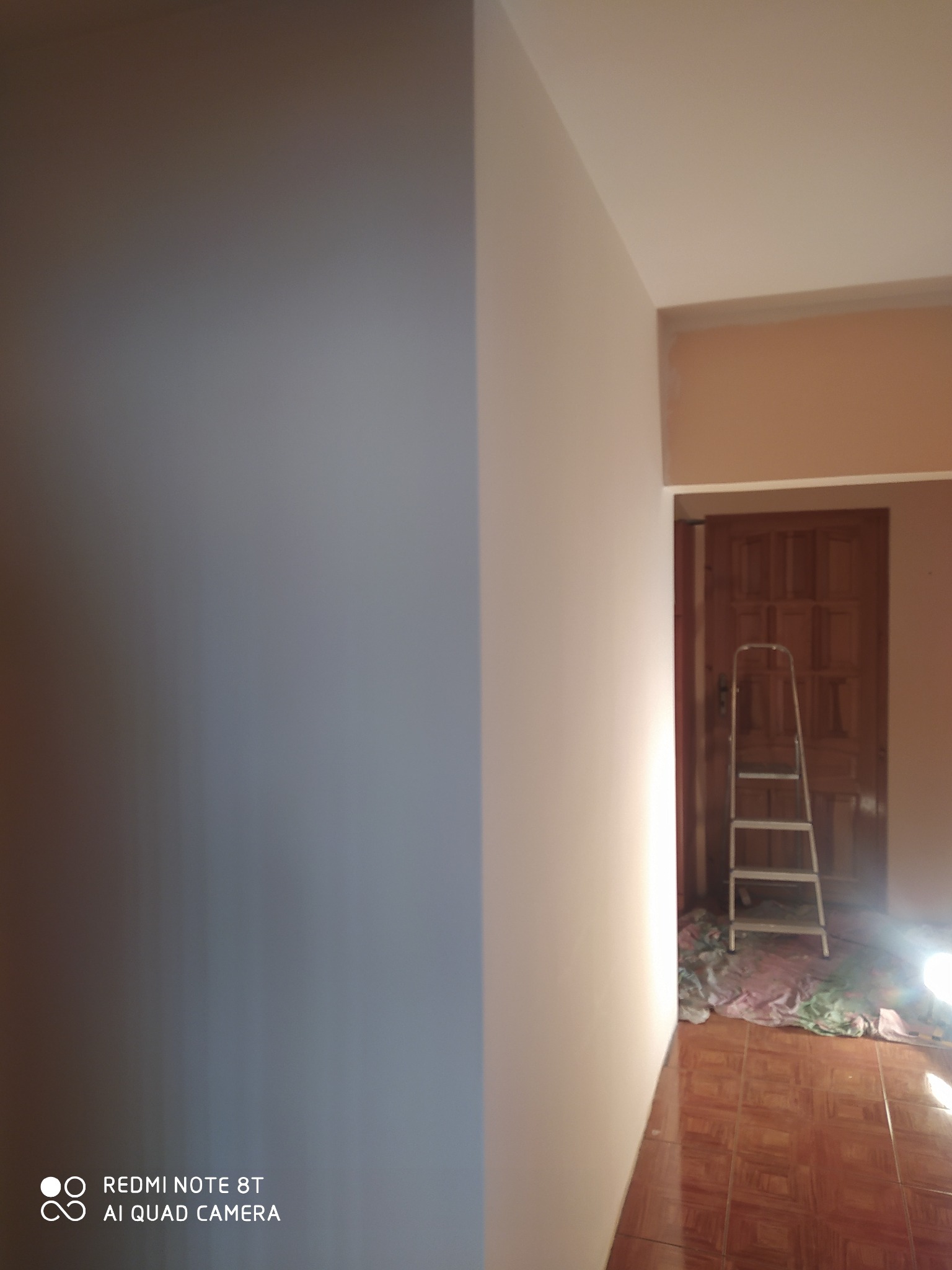 Świeżo pomalowana ściana w odcieniach szarości, widoczna drabina i zabezpieczona podłoga w tle, sugerujące prace wykończeniowe w trakcie realizacji.