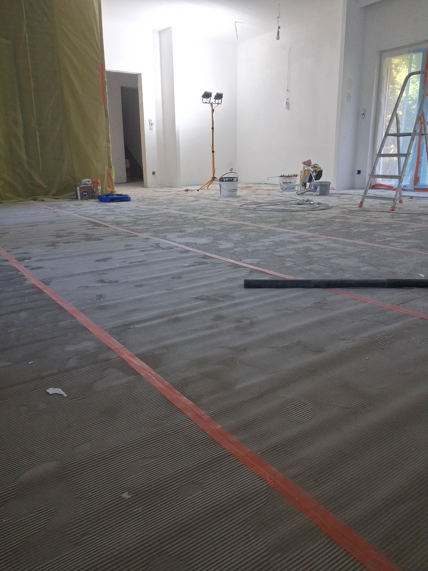 Przygotowanie podłoża pod panele: wylewka samopoziomująca z taśmami dylatacyjnymi w przestronnym, jasnym wnętrzu. Widoczne narzędzia i drabina.
