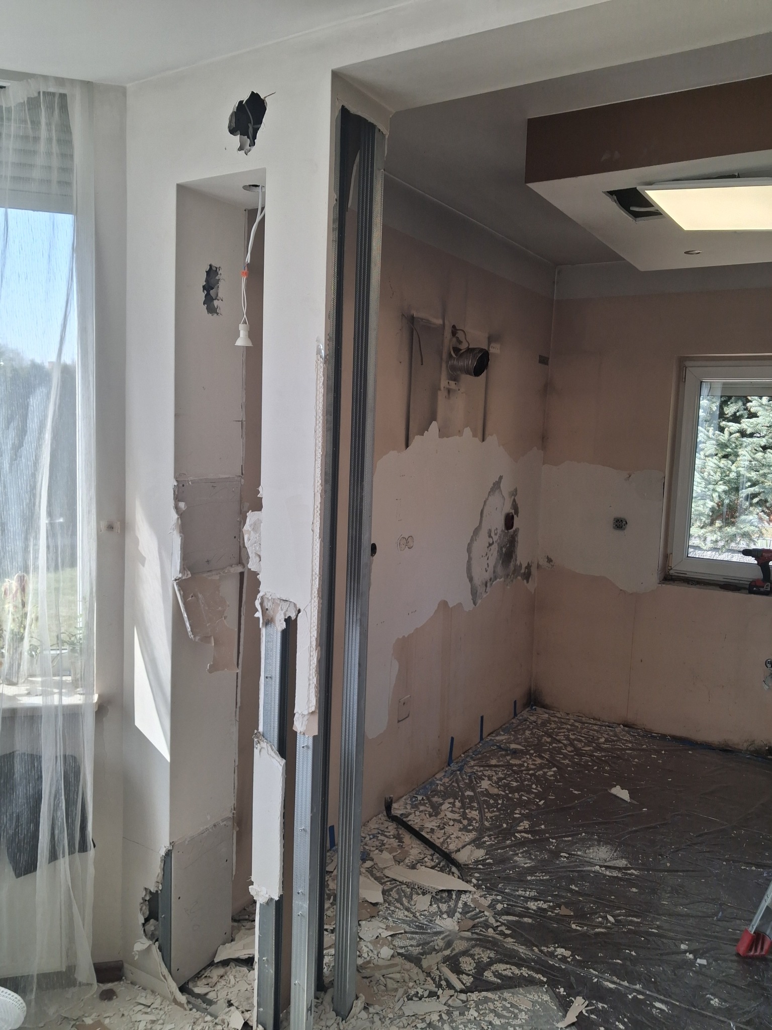 Remont pokoju: odsłonięte ściany z fragmentami gipsu, metalowe profile, zabezpieczona folią podłoga, okno z firanką. Widoczne ślady po instalacjach.