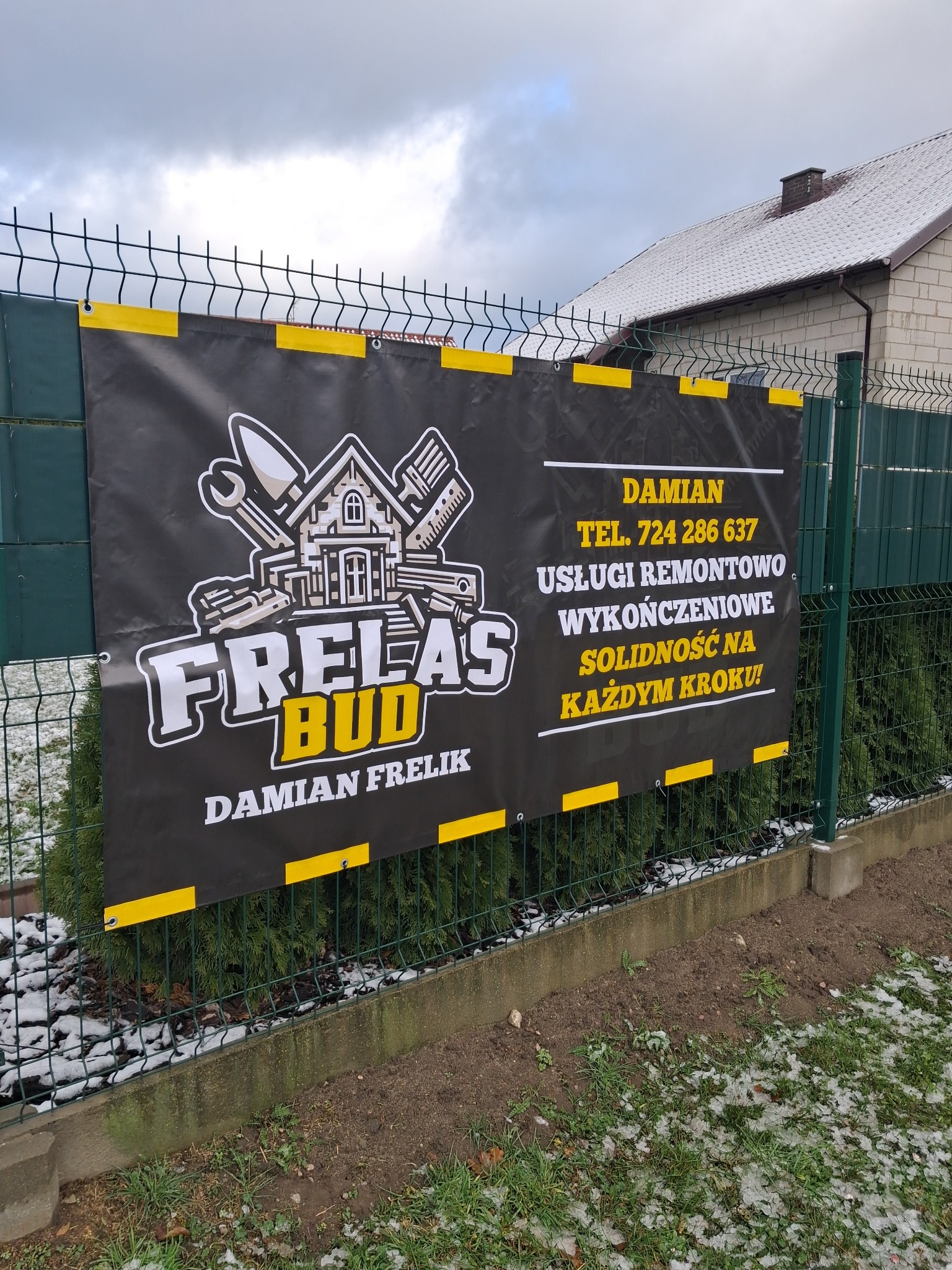 Baner reklamowy firmy remontowo-wykończeniowej 'Frelas Bud' Damiana Frelika, zawieszony na zielonym ogrodzeniu siatkowym. W tle budynek z dachem pokrytym śniegiem.